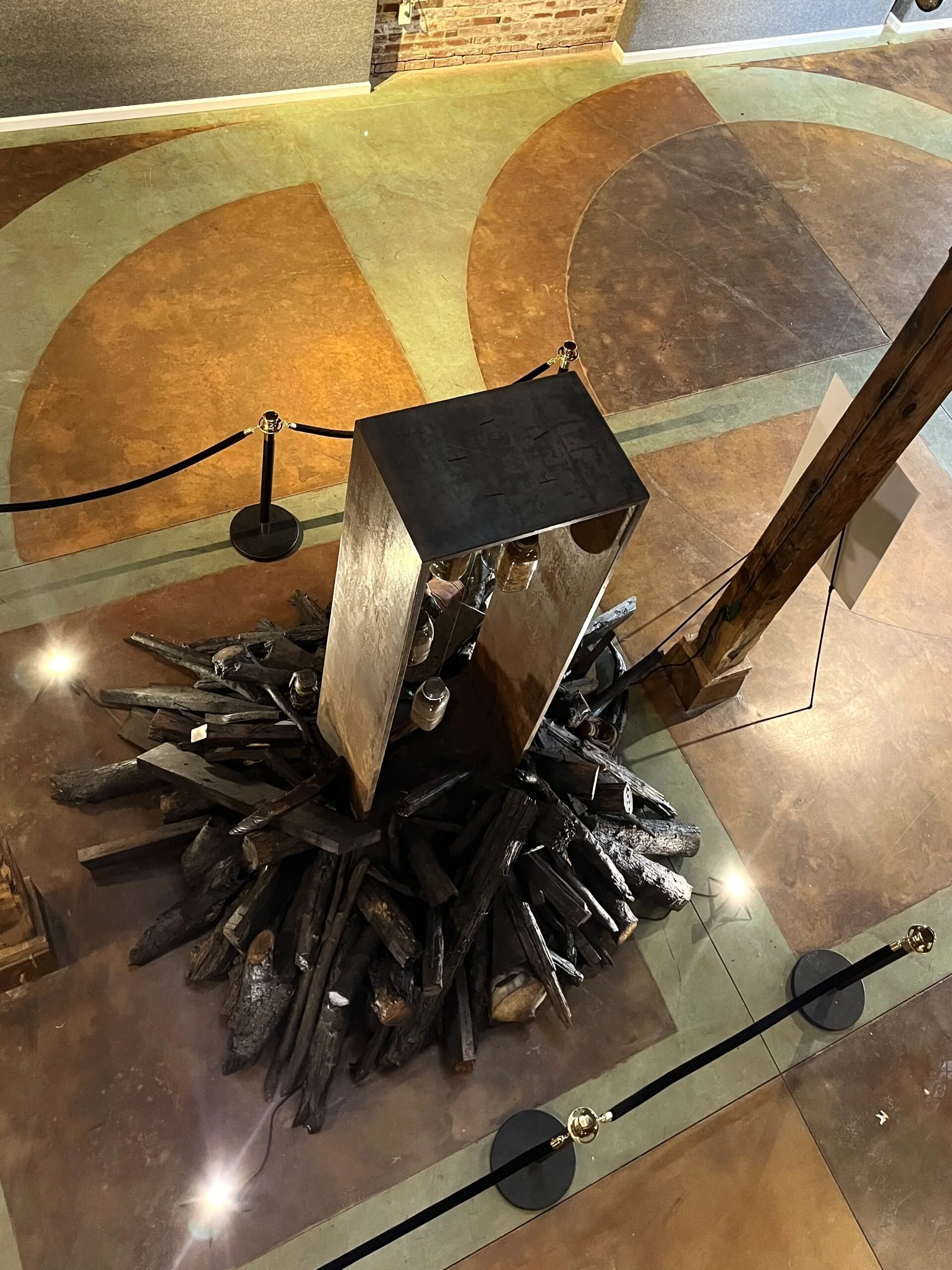 A display of a large pile of charred logs arranged in a circular pattern on a wooden platform in a museum exhibit. The exhibit is roped off with black stanchions and gold accents, with spotlights illuminating the display.