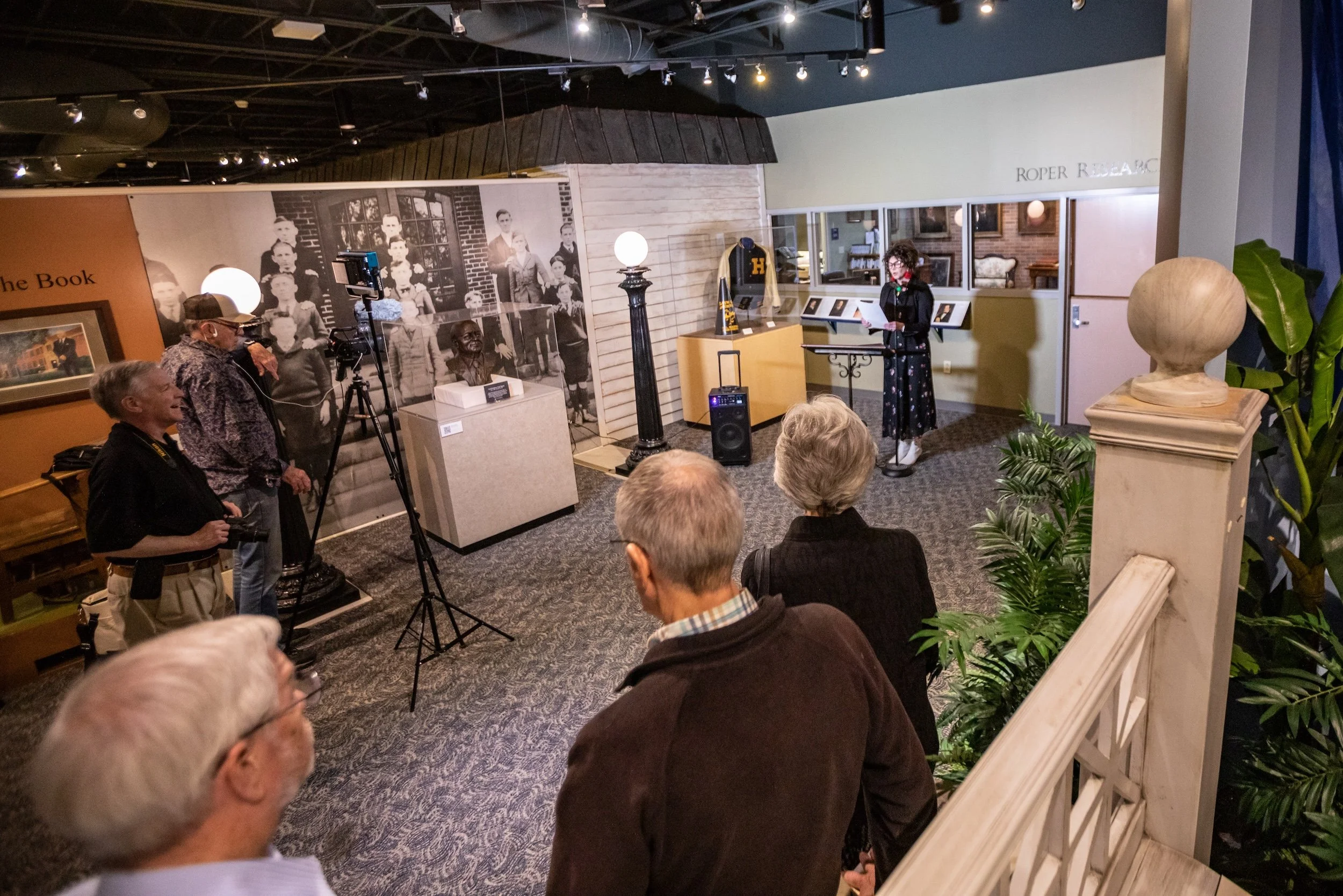 A woman with curly hair and glasses reading from paper at a podium in a museum or exhibit space, with several people seated and watching, and a camera crew recording the event.