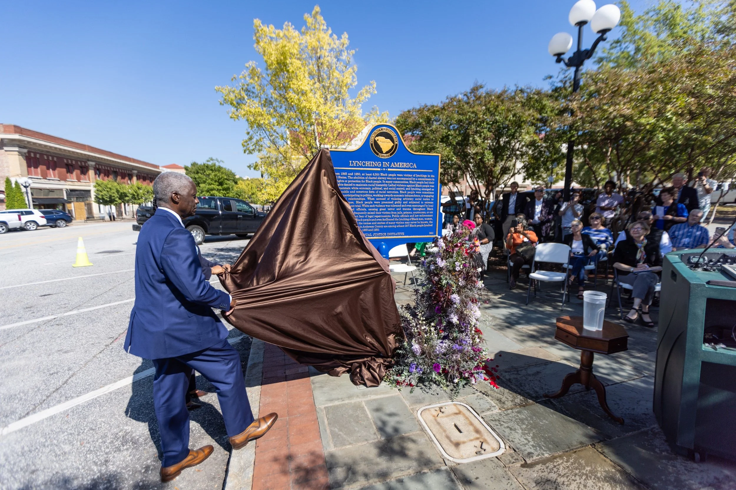 A man in a blue suit unveiling a public historical marker titled 'Lynching in America' during a outdoor event with seated attendees and onlookers, with trees and a building in the background.