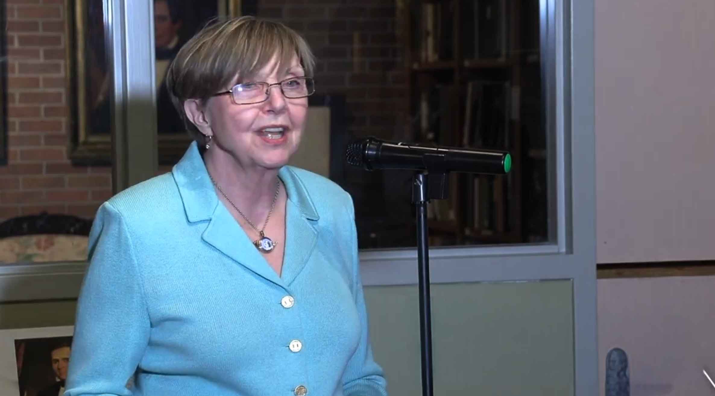 A woman with short light brown hair, glasses, wearing a light blue blazer with pearl buttons, speaking into a microphone indoors with a brick wall and window in the background.