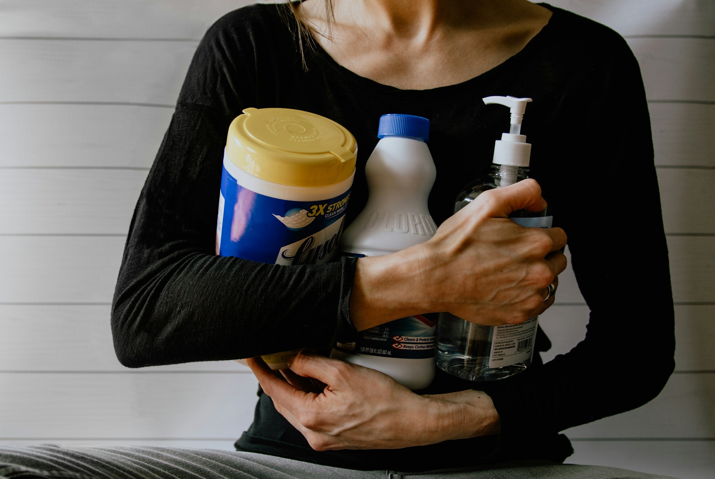 Person holding various household cleaning and hygiene products, including spray bottles and a container of disinfecting wipes, against a light-colored wall with horizontal paneling.