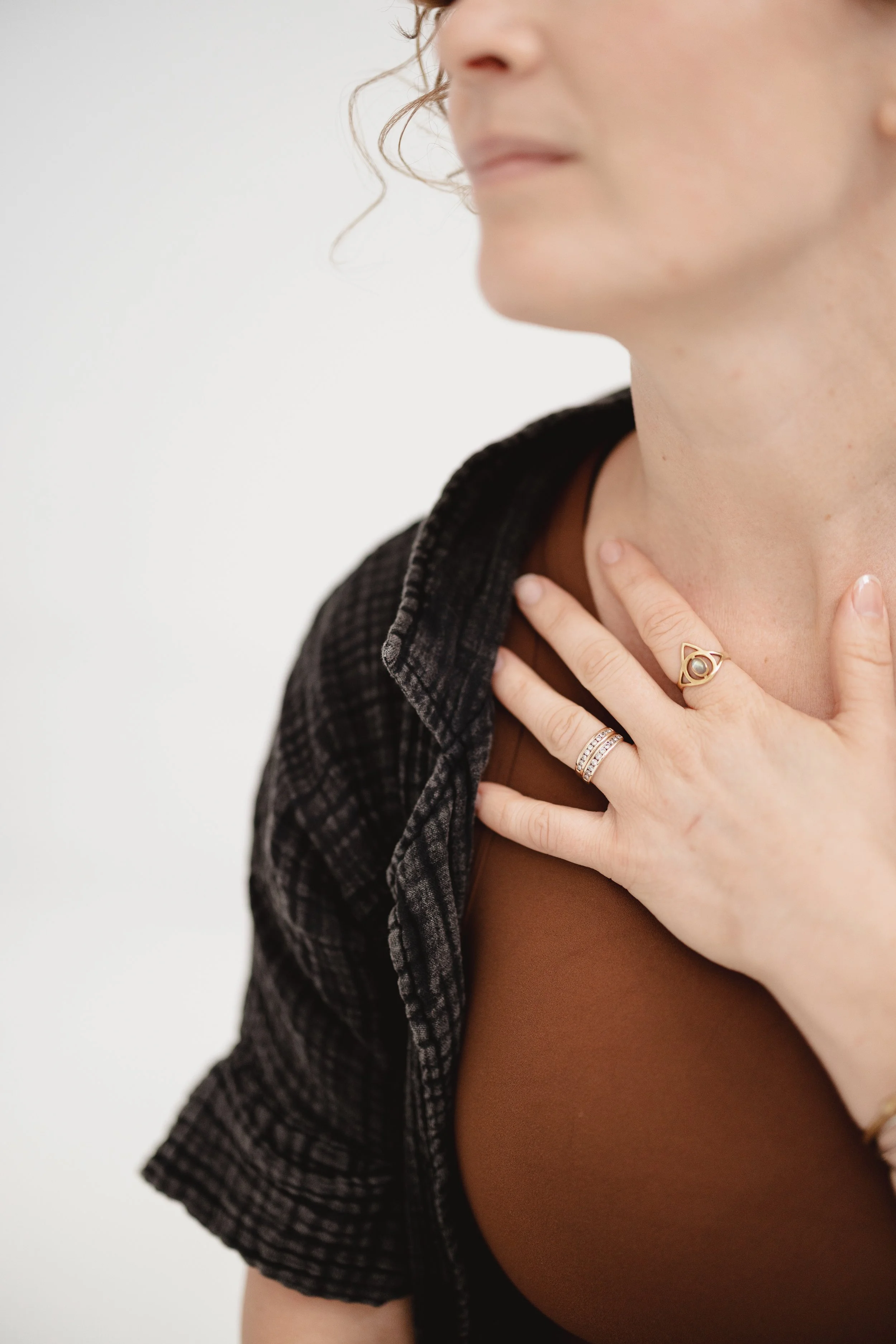 Close-up of a woman's face and hand, wearing rings and a ring on her thumb, with curly hair, touching her chest against a plain white background.