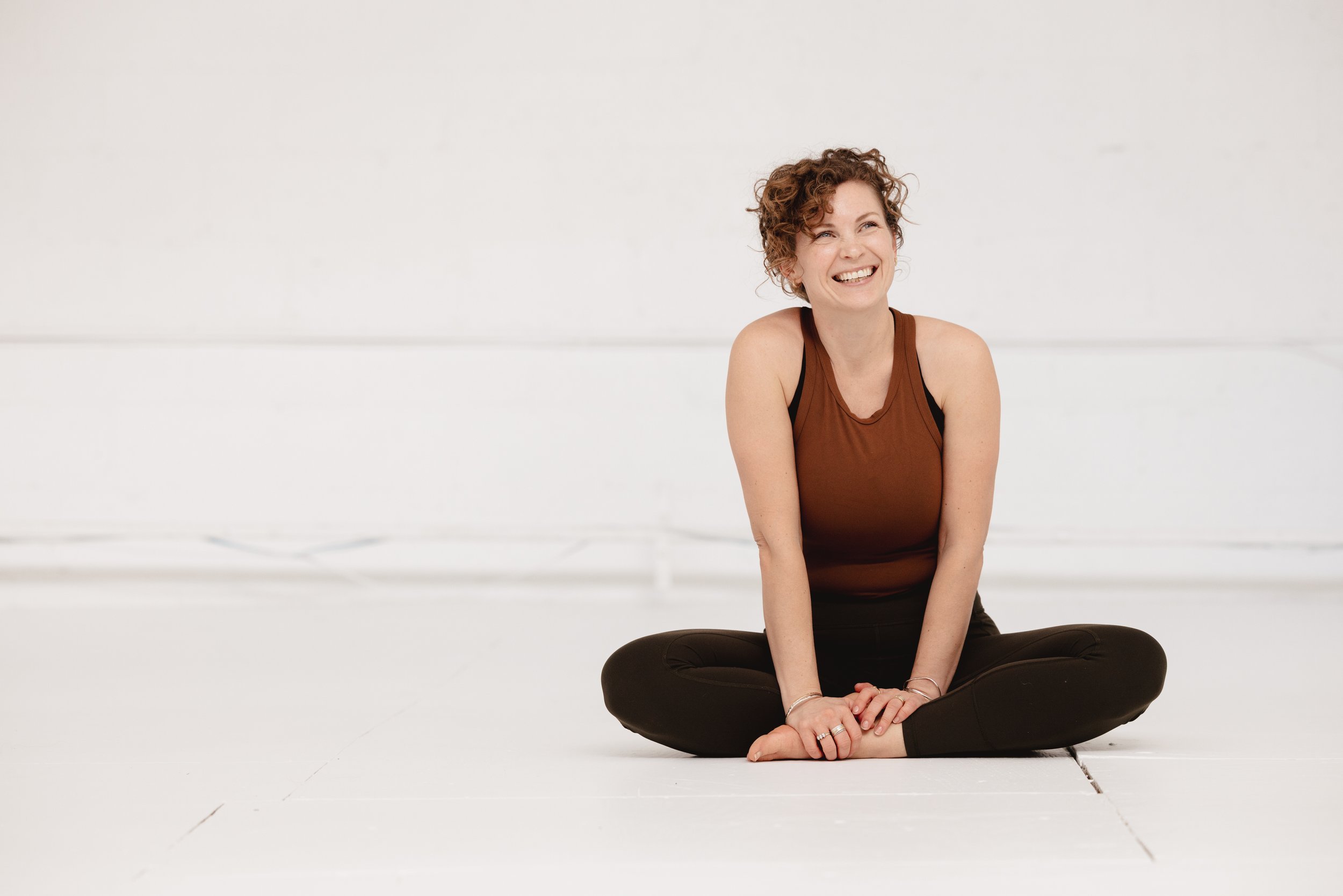 A woman with pulled back curly hair smiling, sitting cross-legged on the floor in a minimalistic white room.