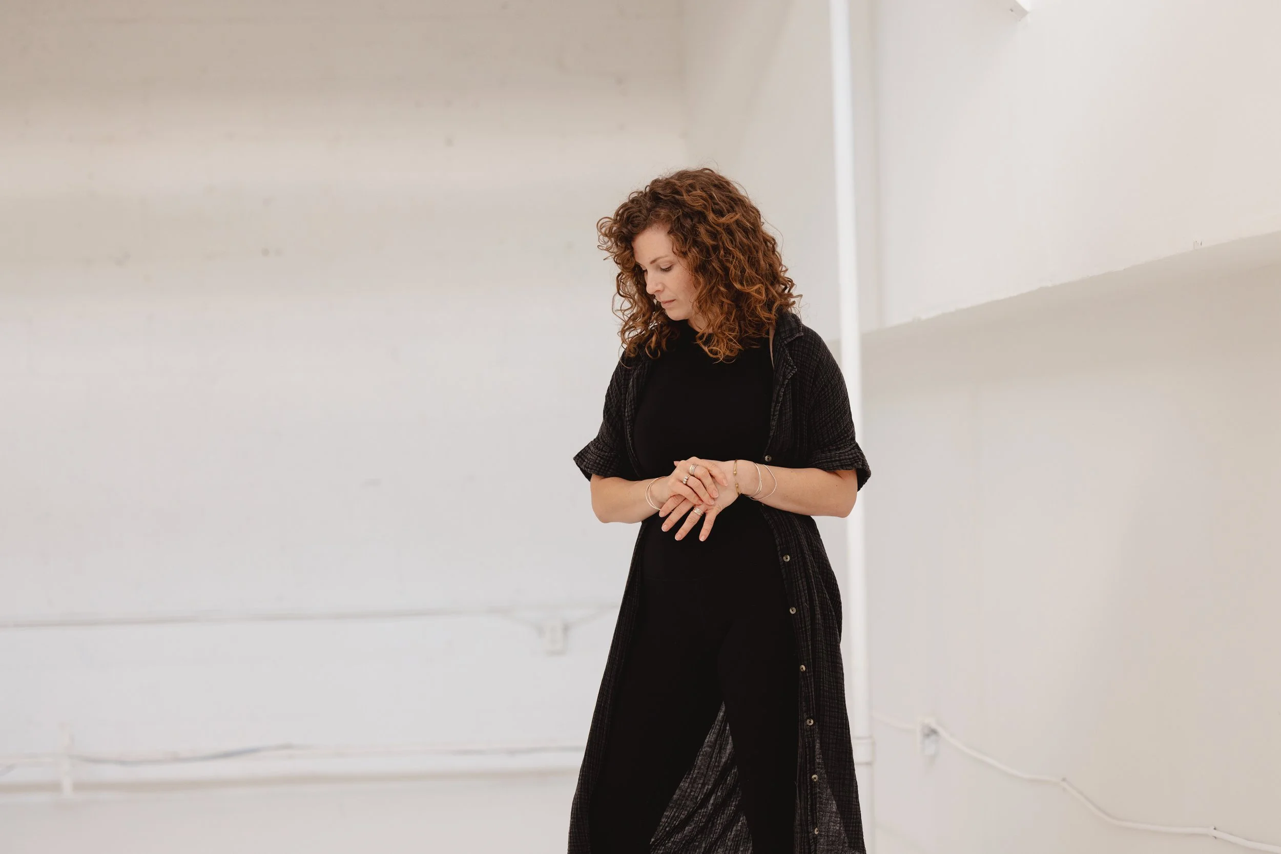 A woman with curly red hair wearing a black top and a sheer black cover-up, standing in front of a white wall with her head tilted downward and arms crossed, in a minimalistic, brightly lit room.