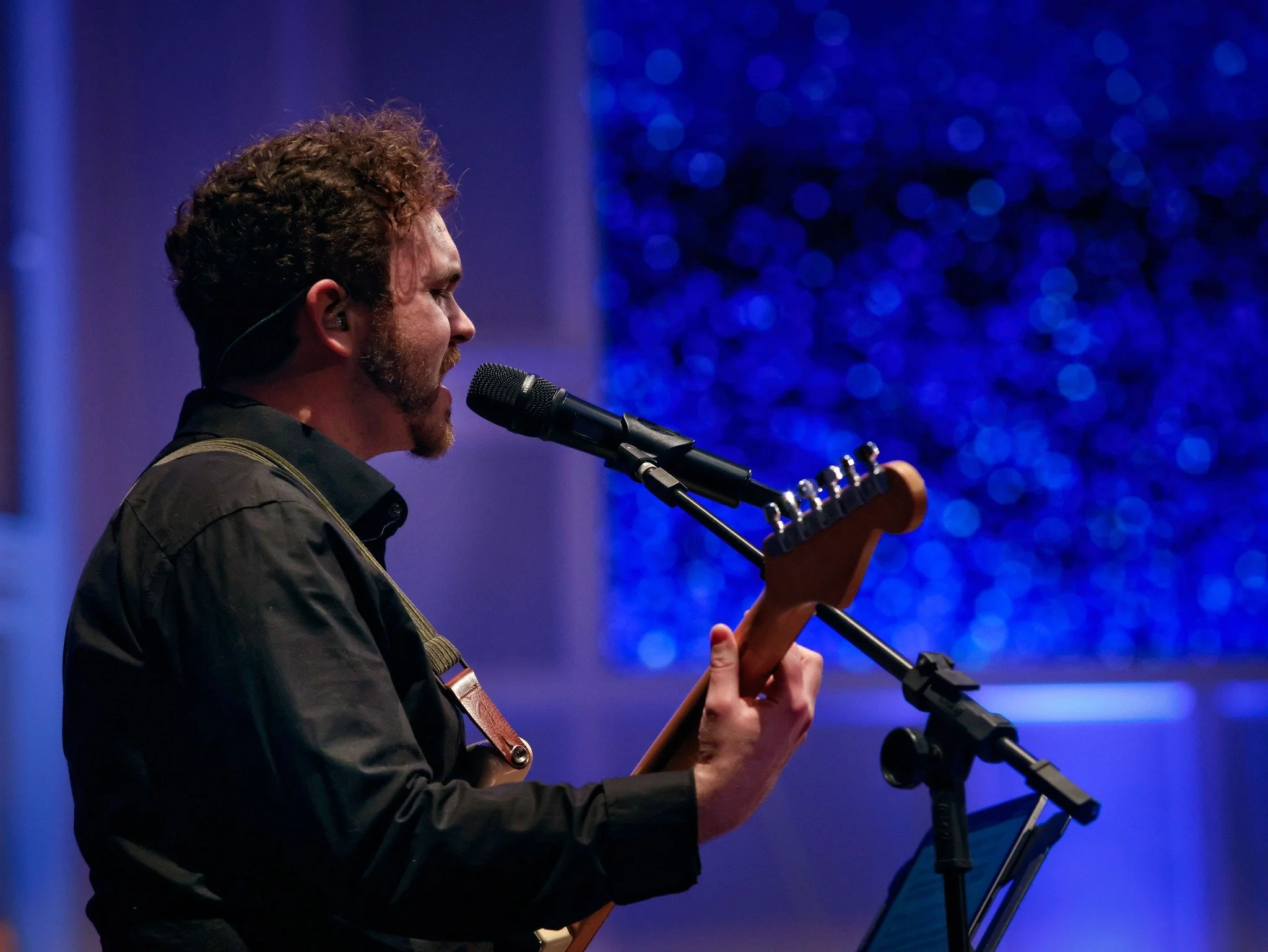 A man with curly hair and a beard playing an electric guitar and singing into a microphone on stage with a blue-lit background.