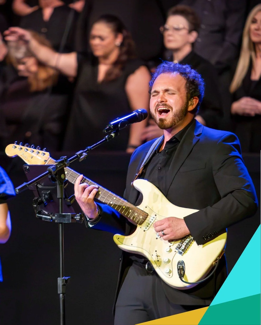 A male singer and guitarist passionately performing on stage with eyes closed. He is playing an electric guitar in front of a choir or backup singers, who are standing behind him and singing.