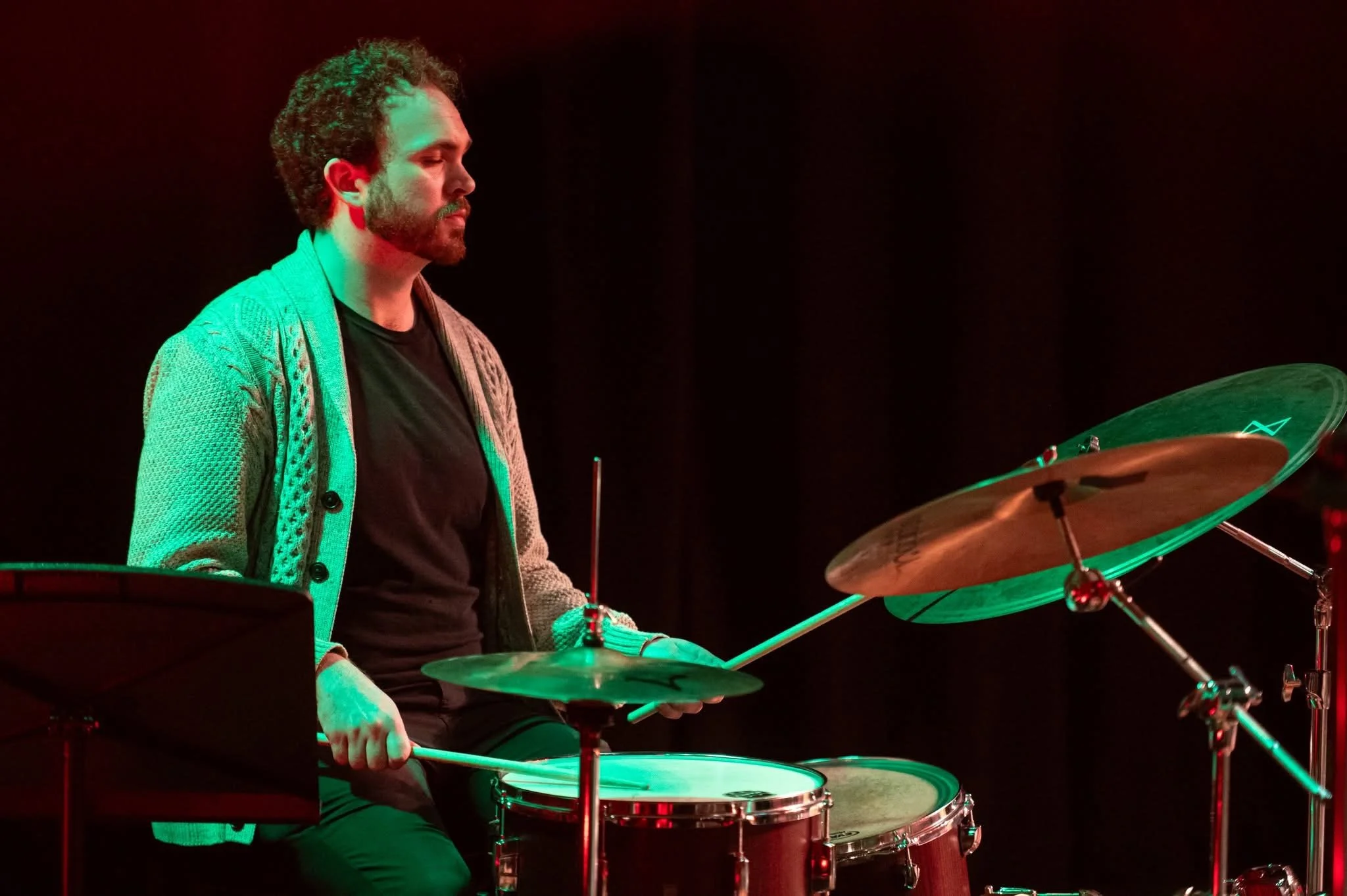 A man with curly hair and a beard playing drums on a stage with red and green lighting.
