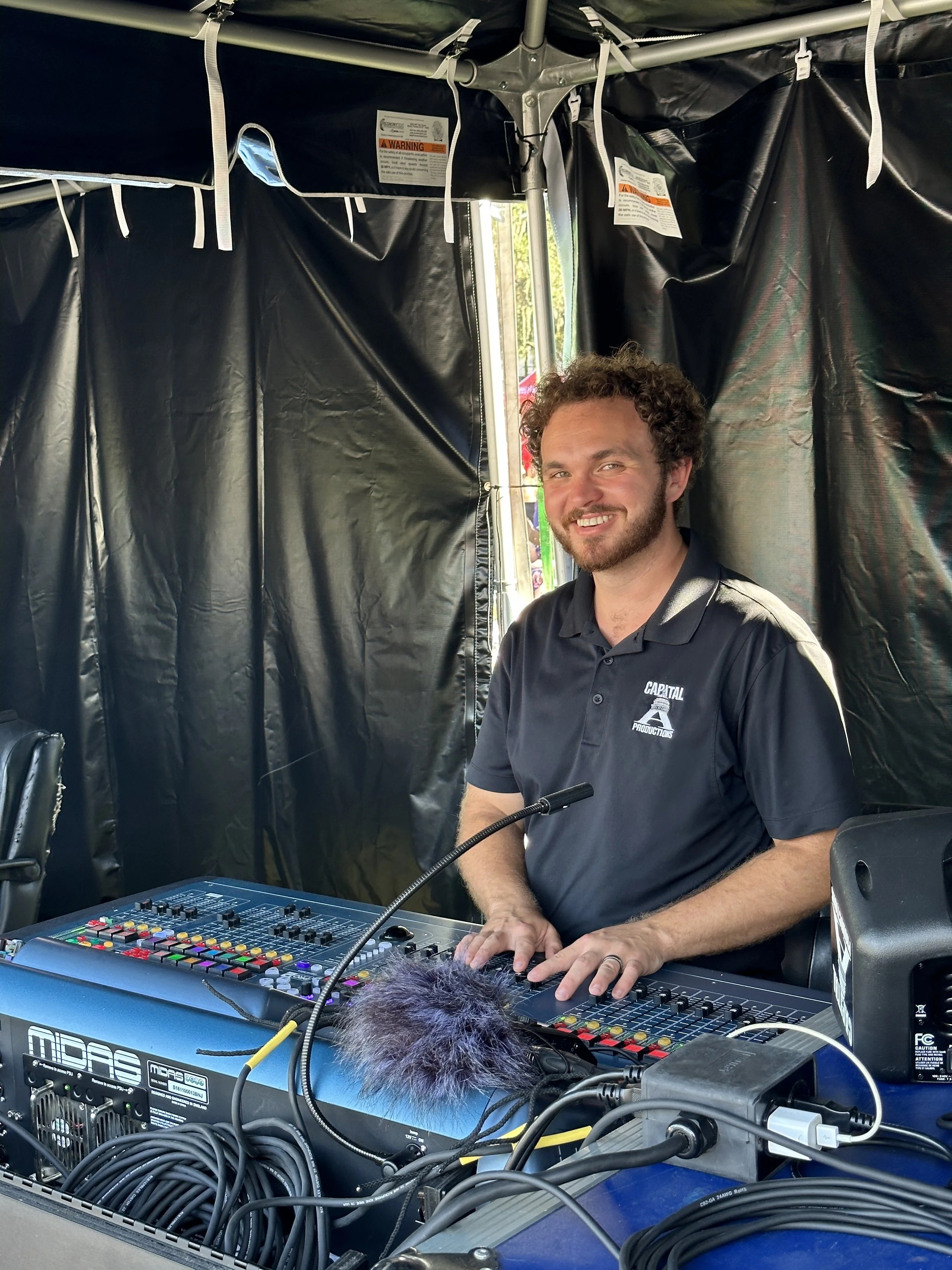 A man with curly hair and a beard smiling while operating sound equipment at an event. He is sitting behind a sound mixer and the setting appears to be a black tent.