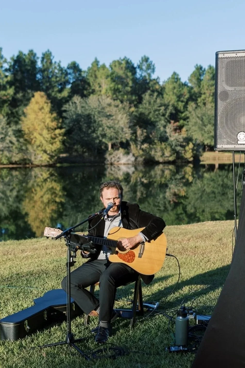 A man playing an acoustic guitar and singing into a microphone outdoors near a river with green trees in the background.