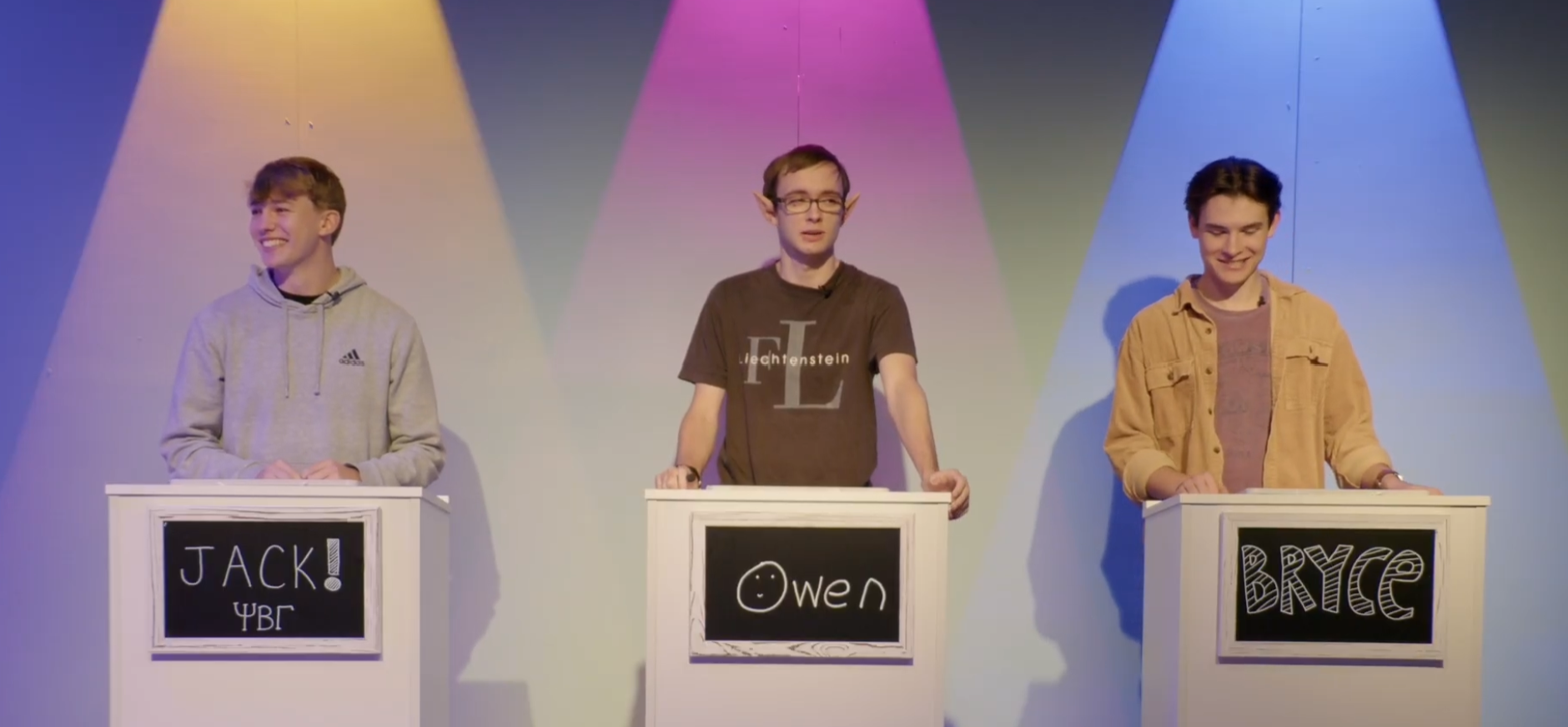 Three young men standing behind podiums with blackboards displaying their names, Jack, Owen, and Bryce, on a colorful stage with multicolored spotlights overhead, participating in a quiz show.