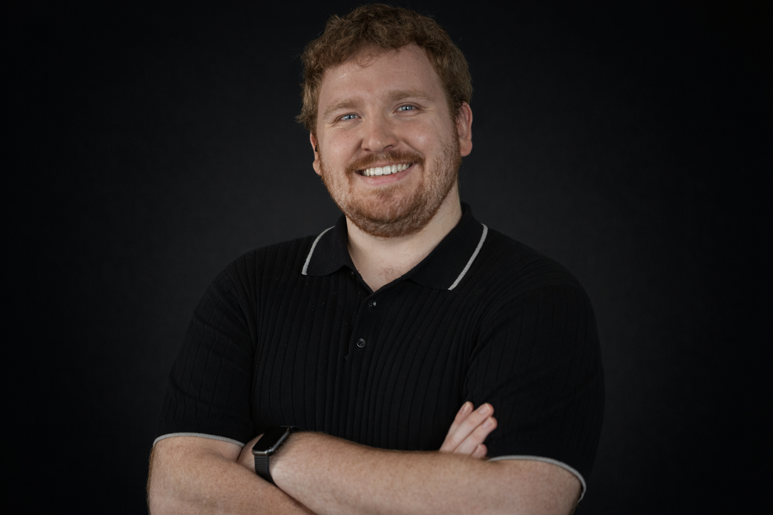 A smiling man with curly red hair, beard, and blue eyes, wearing a black polo shirt with white trim, and a smartwatch on his left wrist, standing against a dark background.