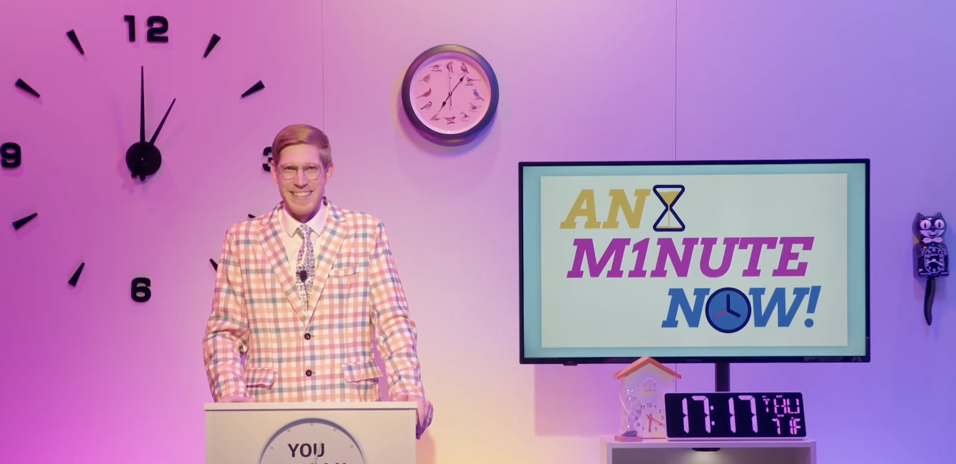 A man wearing glasses and a checkered blazer standing in front of a colorful set with clocks and a sign that reads 'An Minute Now!' with a clock icon. The background is pink and purple with a large wall clock and a smaller analog clock above him, and a digital clock displaying 17:17:04.