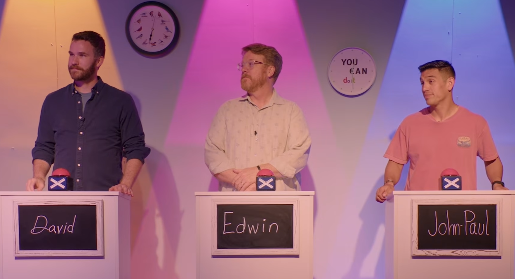Three men standing behind podiums with red buzzer buttons on a game show set. The left man has a beard and dark shirt, the middle man wears glasses and a light shirt, and the right man has short hair and a pink T-shirt. Behind them, a clock and a sign that says "You Can do it" are visible.