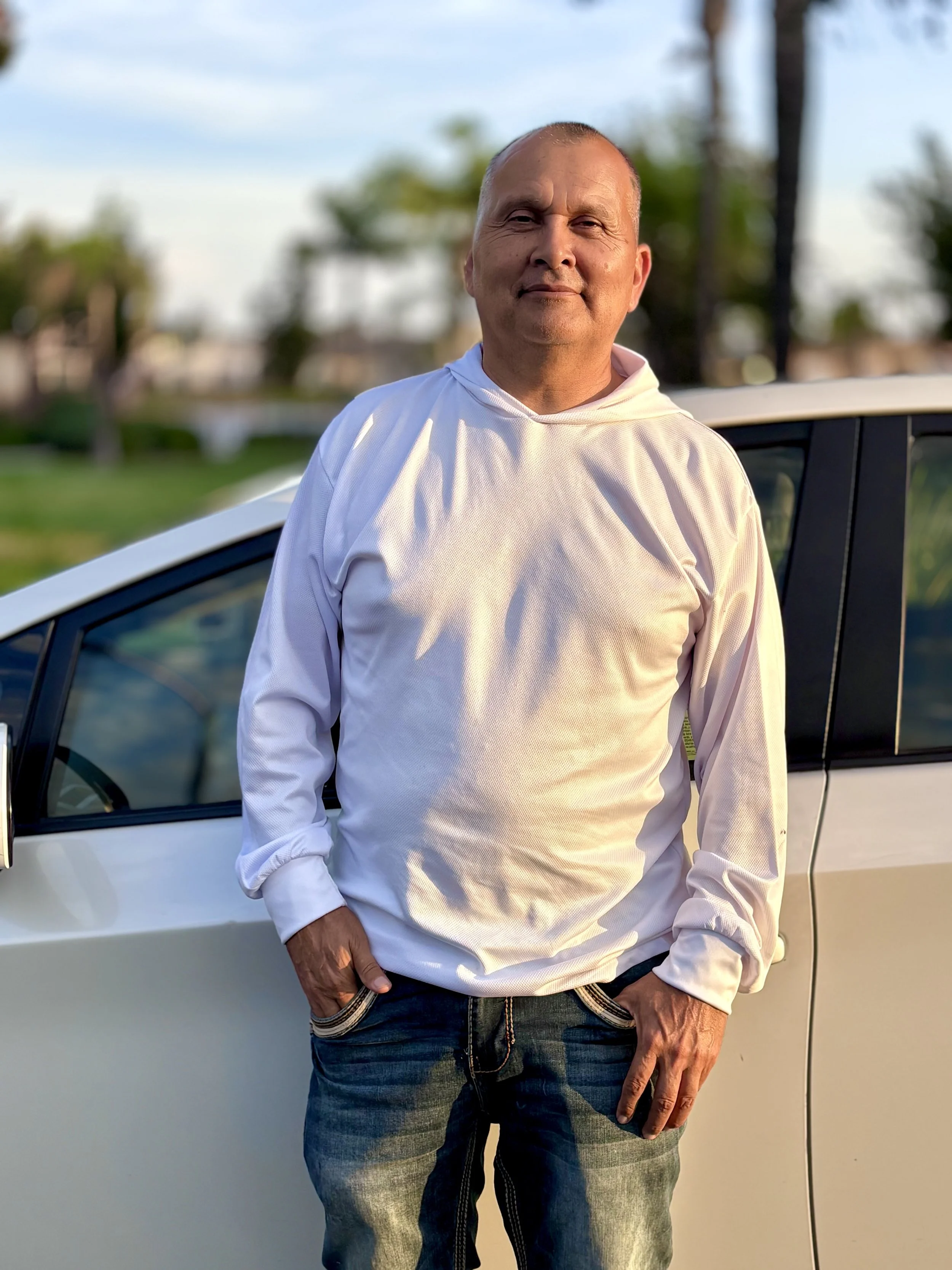 A man in a white hoodie and jeans standing in front of a white car, with palm trees and a partly cloudy sky in the background during daylight.