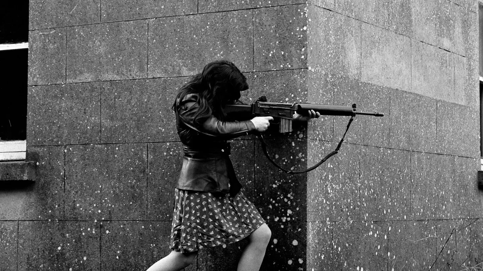 A woman with long hair, wearing a leather jacket and a patterned skirt, aiming a rifle while leaning against a speckled concrete wall.