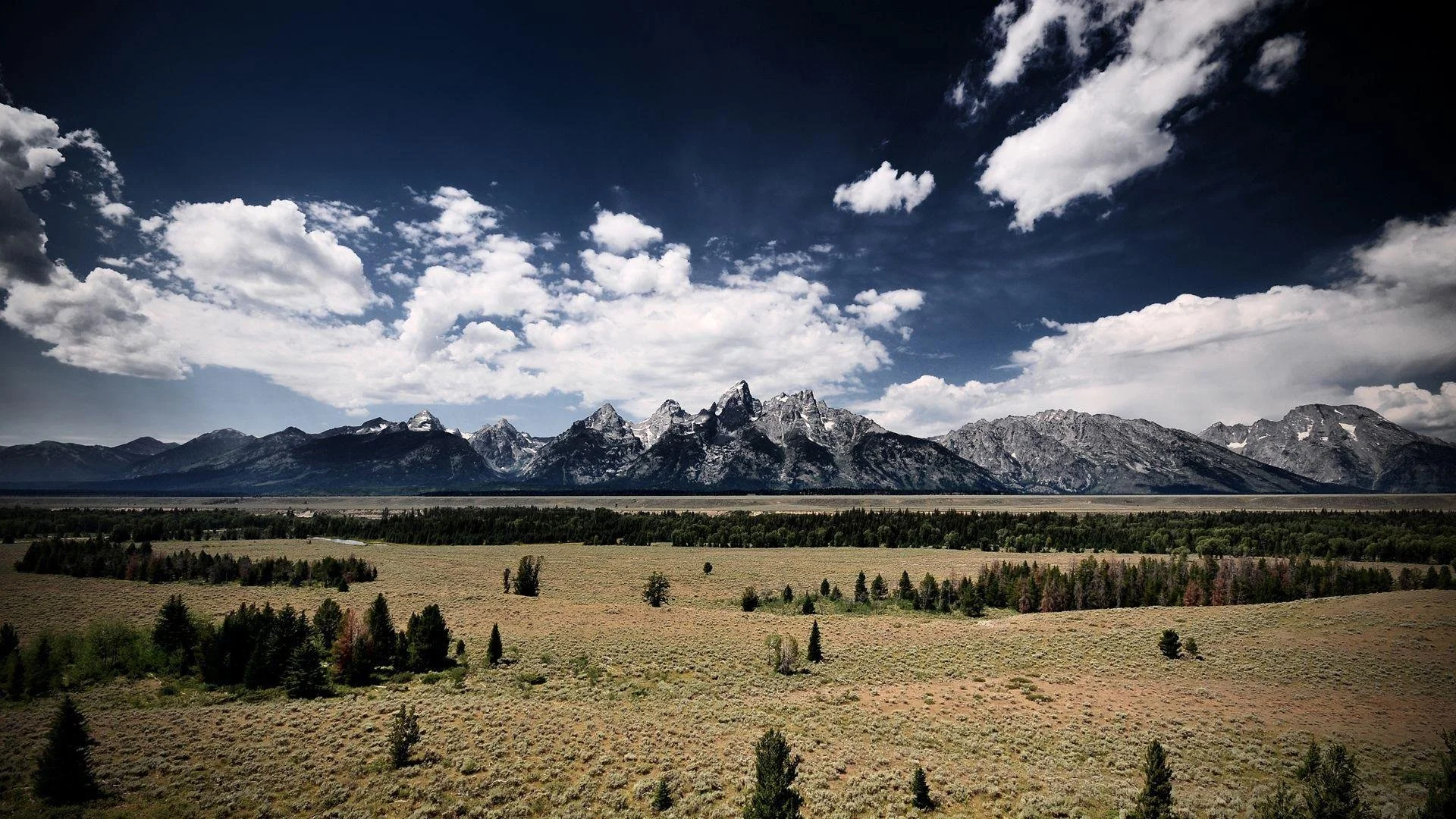 Mountain range with snow-capped peaks under a partly cloudy sky, foreground features forested plains.