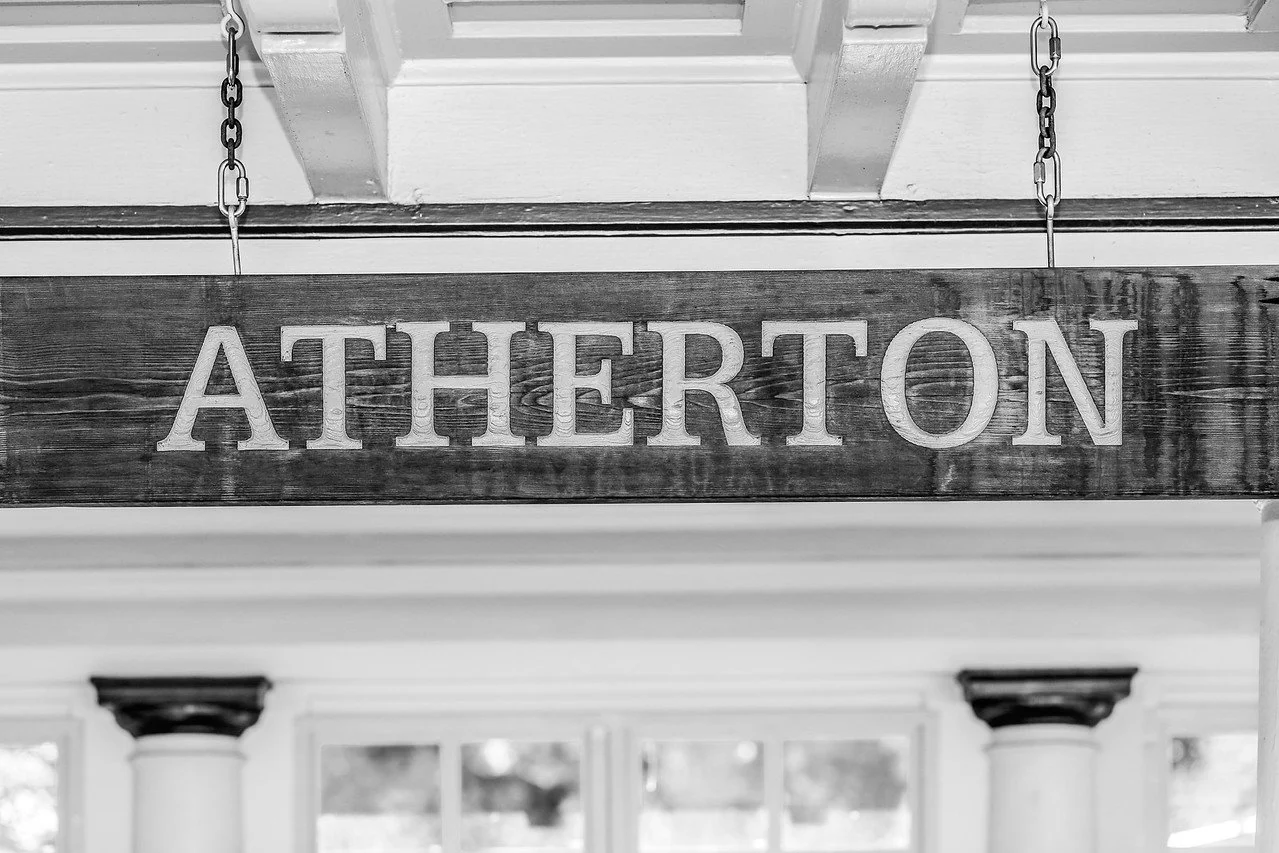 A wooden sign with the word 'ATHERTON' hanging from the ceiling by chains inside a house or building.