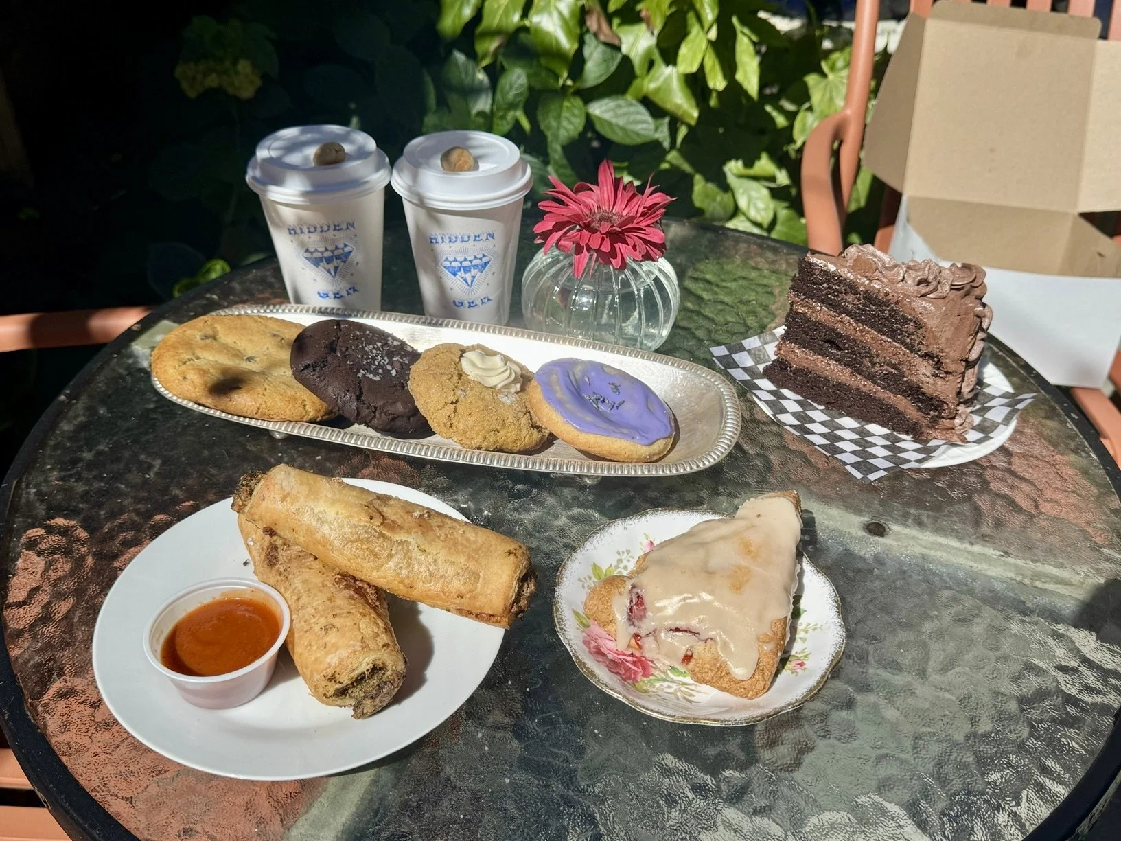 A round table with assorted desserts including a slice of chocolate cake, cookies, and a strawberry scone, with two cups of coffee, two savoury vegan rolls with tomato sauce, and a partially opened box in the background.