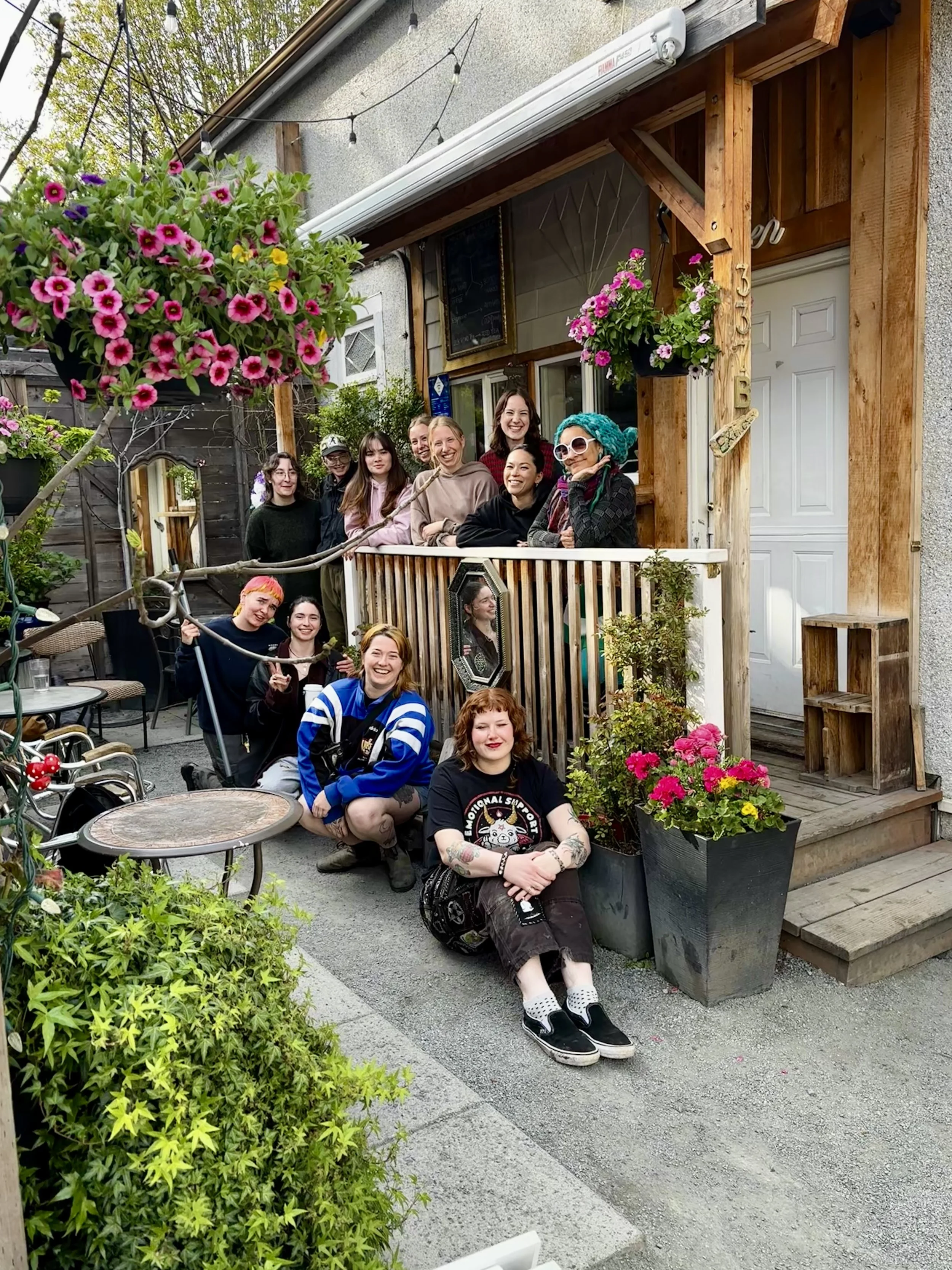 A group of people gathered on the porch and steps of a small building, with potted flowers and greenery around them, smiling and posing for a photo.
