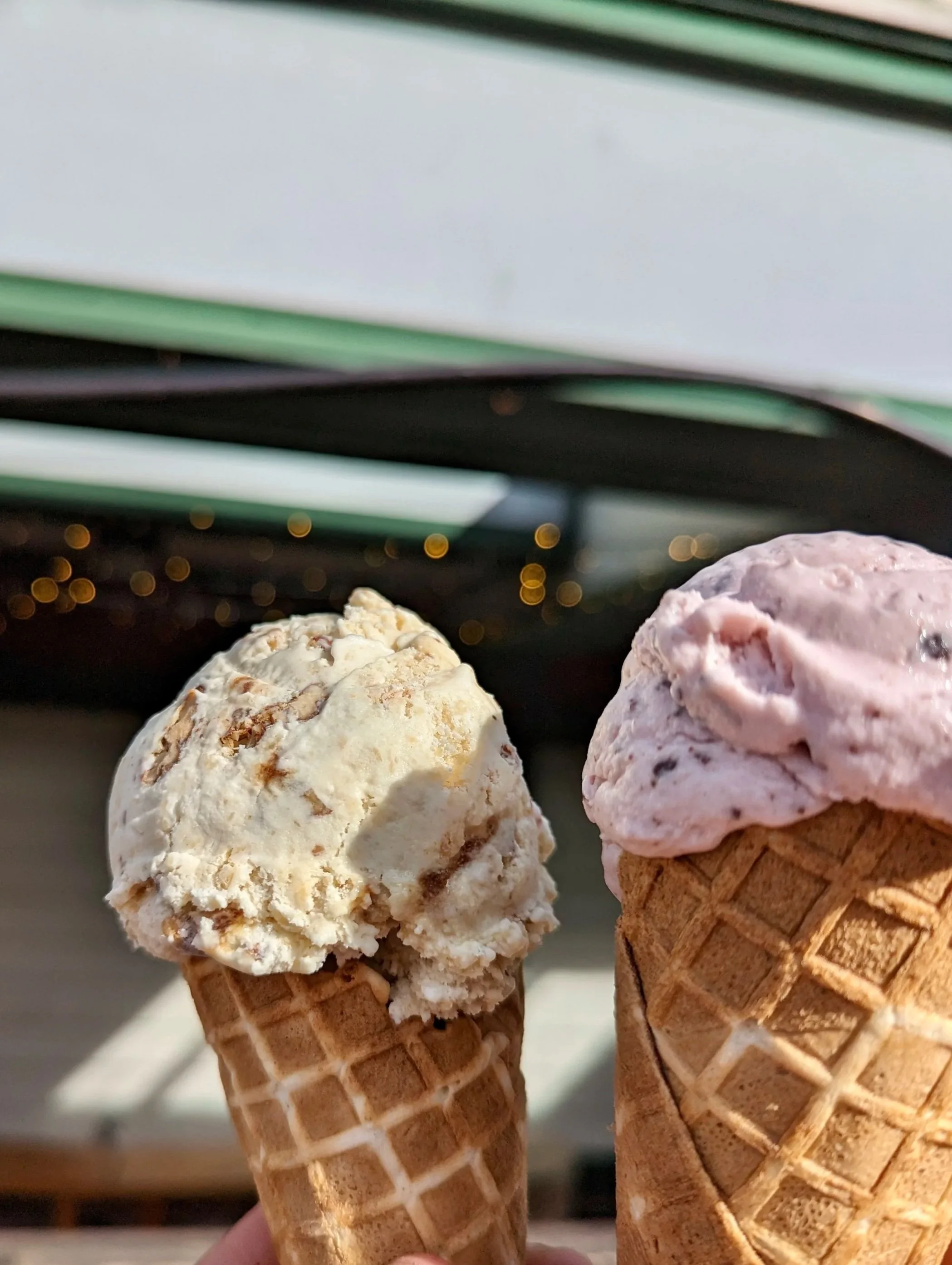 Two ice cream cones with scoops of candied walnut and raspberry chocolate chip in waffle cones, held outdoors with blurred background and lights.