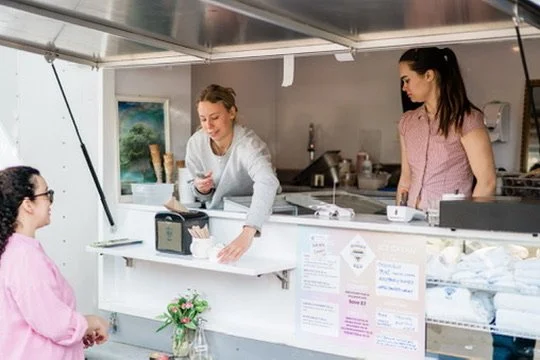 A woman at a food truck counter serving ice cream, with another woman working inside the truck in the background.