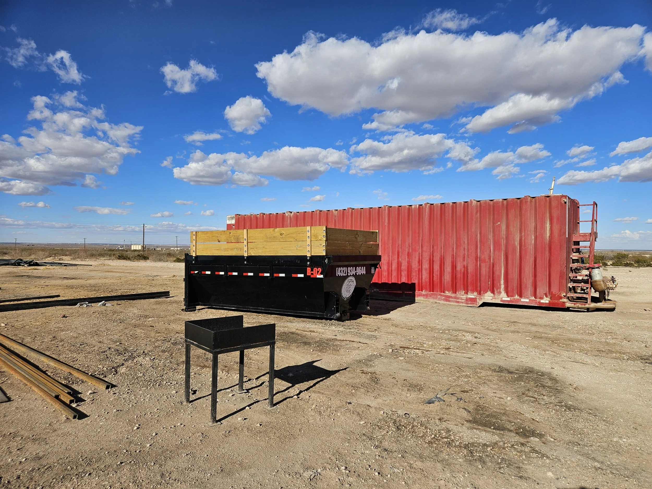 A barren landscape with train tracks in the foreground, a red shipping container on a flatbed trailer, wooden planks, and a blue sky with scattered white clouds.