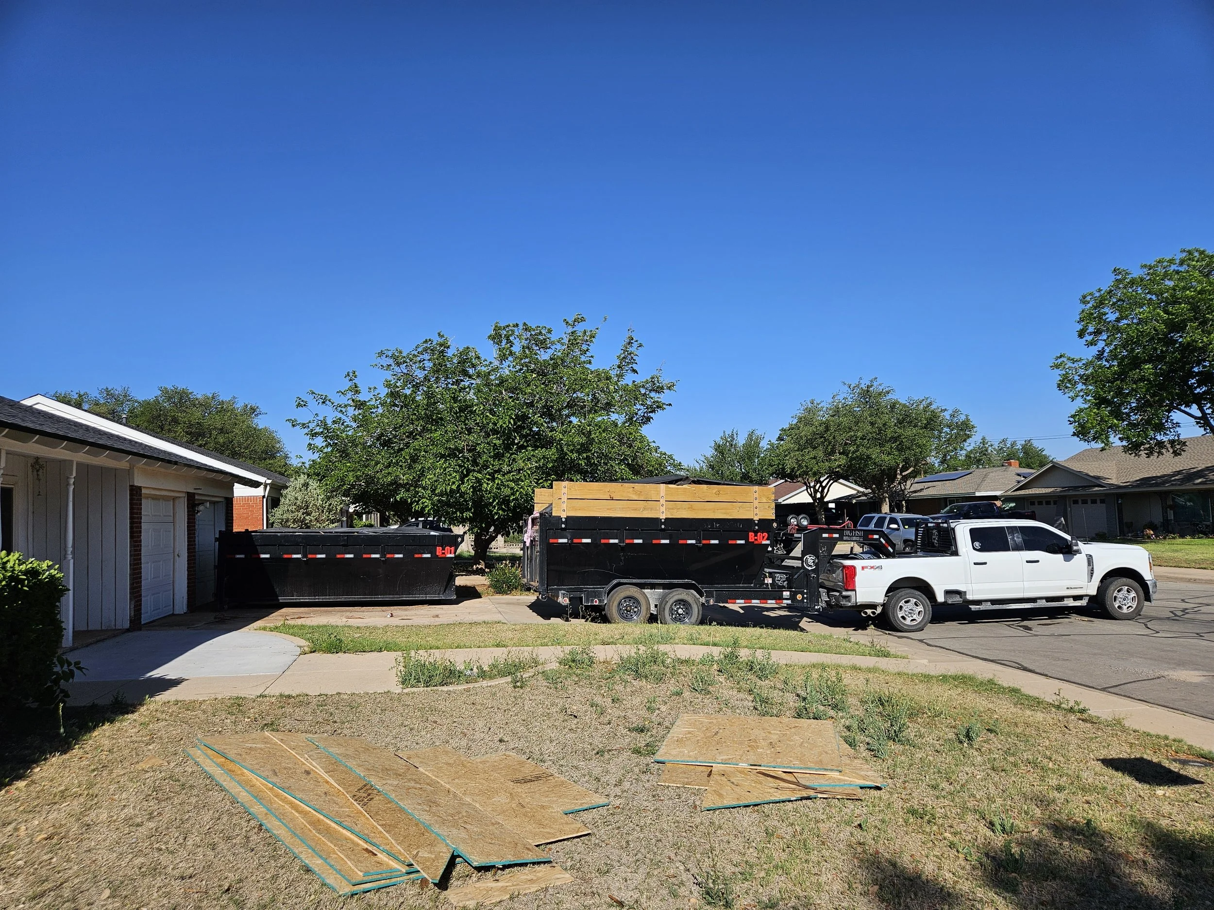 Residential backyard with trees, a driveway with parked trucks, and construction materials on the grass in sunny weather.