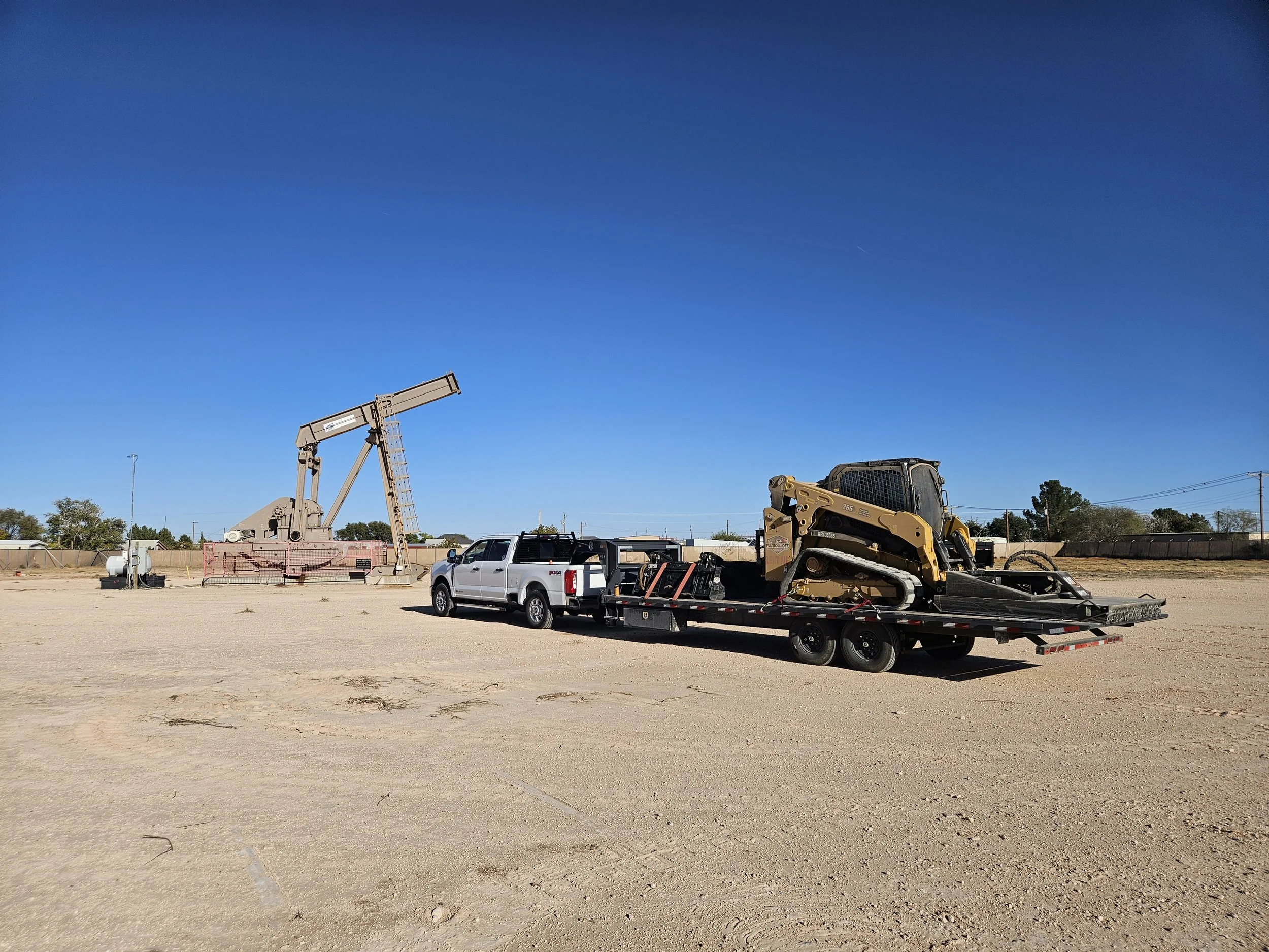 A flatbed trailer carrying construction equipment, including a yellow skid steer loader, at a construction site with a large excavator and pickup truck nearby. The background shows a clear blue sky, some trees, and power lines.