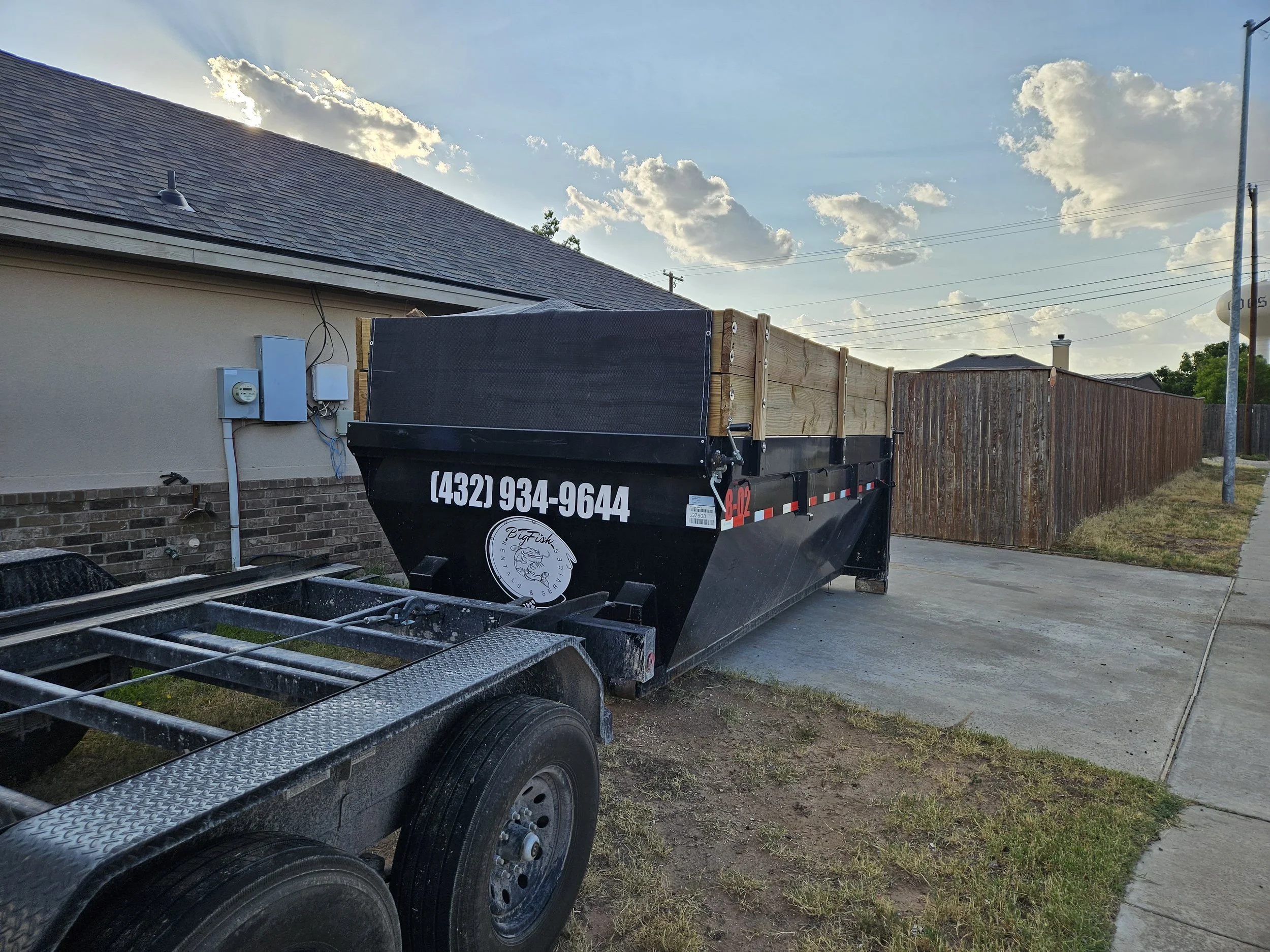 A trailer parked next to a house with a shingled roof, utility meters, and a wooden fence in a suburban neighborhood with a cloudy sky.