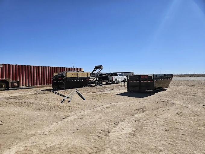 Vehicles and trailers parked on a desert-like open area with a clear blue sky; including a white pickup truck, a small black trailer with stacked wood, and a large black trailer.