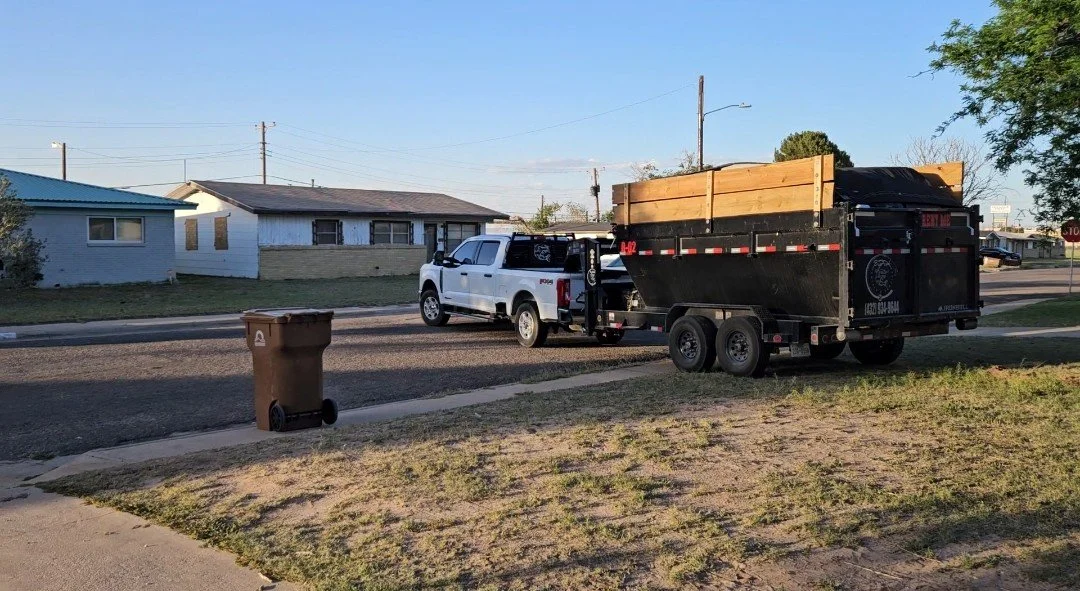 A white pickup truck pulling a black trailer with wooden planks in a residential neighborhood with houses, a trash bin on the sidewalk, and a tree on the right.
