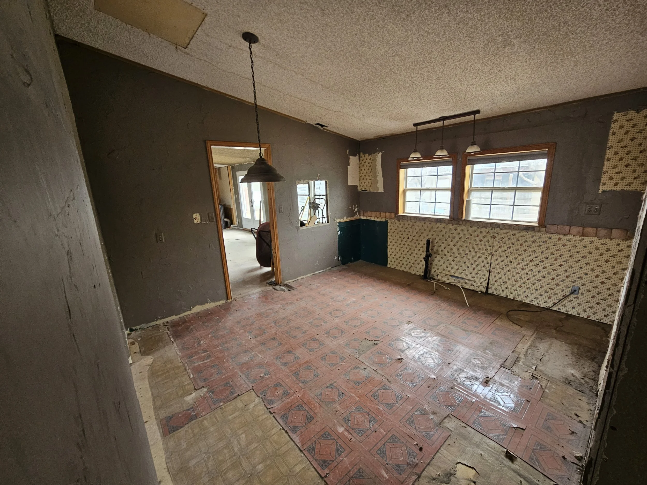 Interior of a room under renovation with partially removed old wallpaper, exposed brick on the walls, and vintage patterned linoleum flooring. There are two large windows letting in natural light, a door opening into another room, and an unfinished c