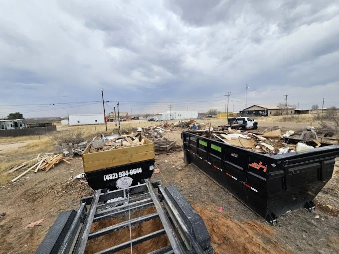 Construction site with two large black dumpsters filled with debris, piles of wood and materials on the ground, and a cloudy sky overhead.