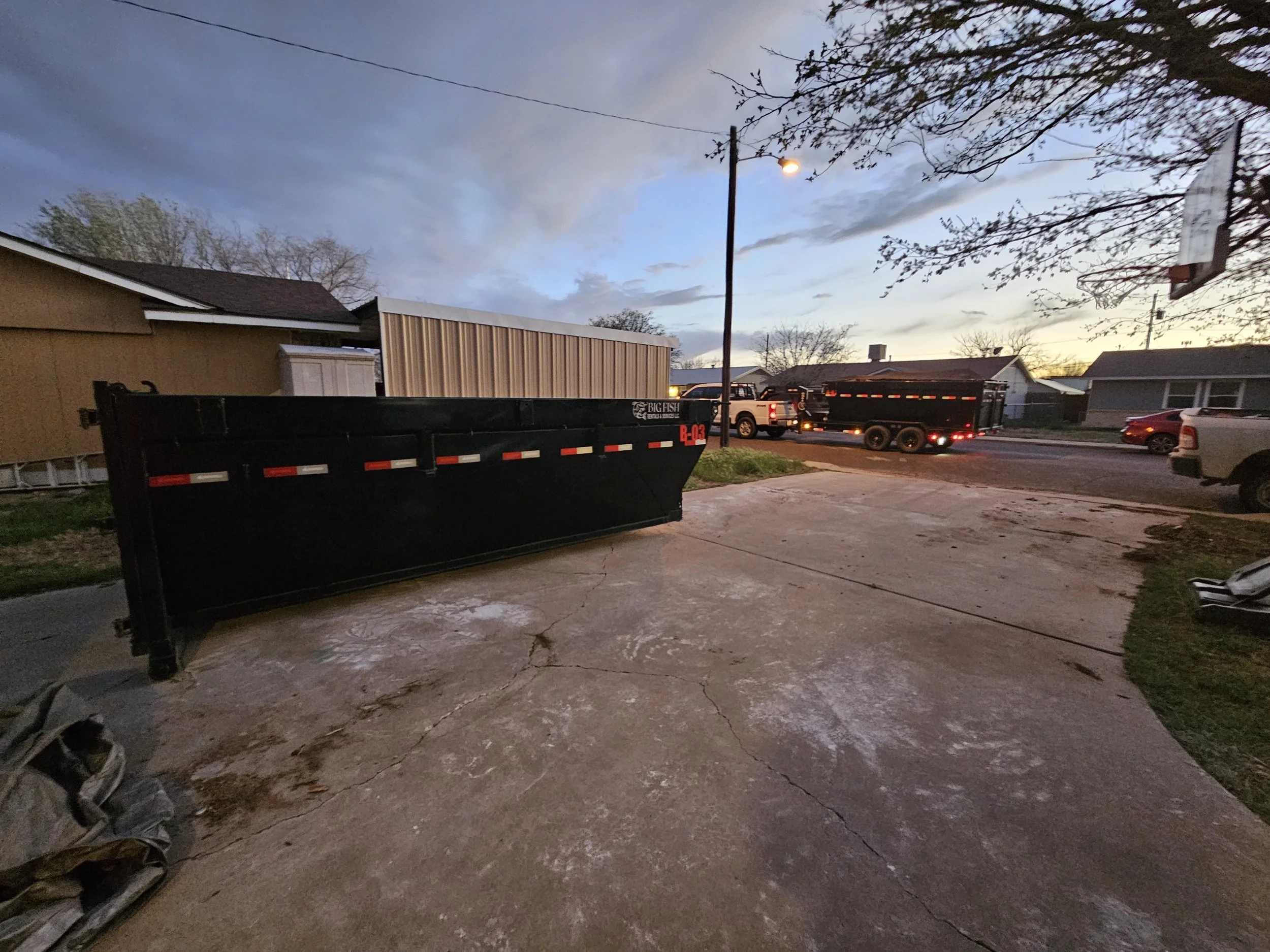 Residential backyard in the early evening, showing a large black dump trailer and a pickup truck on the street, with a basketball hoop attached to a house on the right and a utility pole with wires. The sky is partly cloudy.