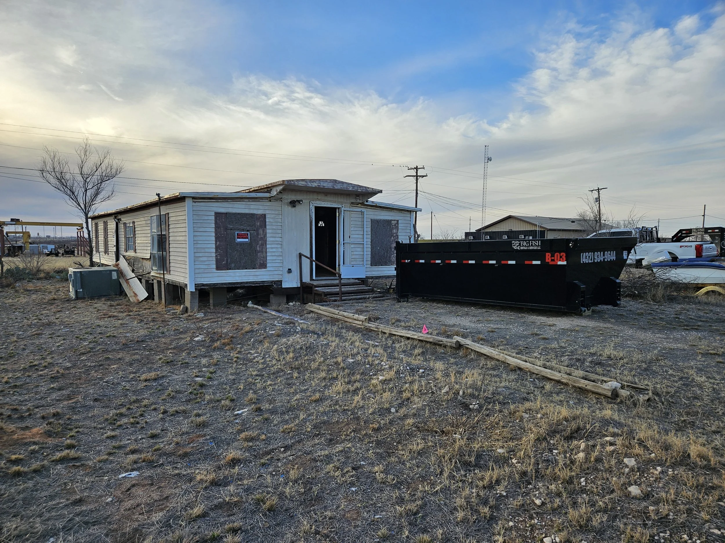 A small white house with boarded-up windows and a partially open front door, situated in an open, grassy area with a tree, utility poles, and a tower in the background. There are also trailers and a boat nearby.
