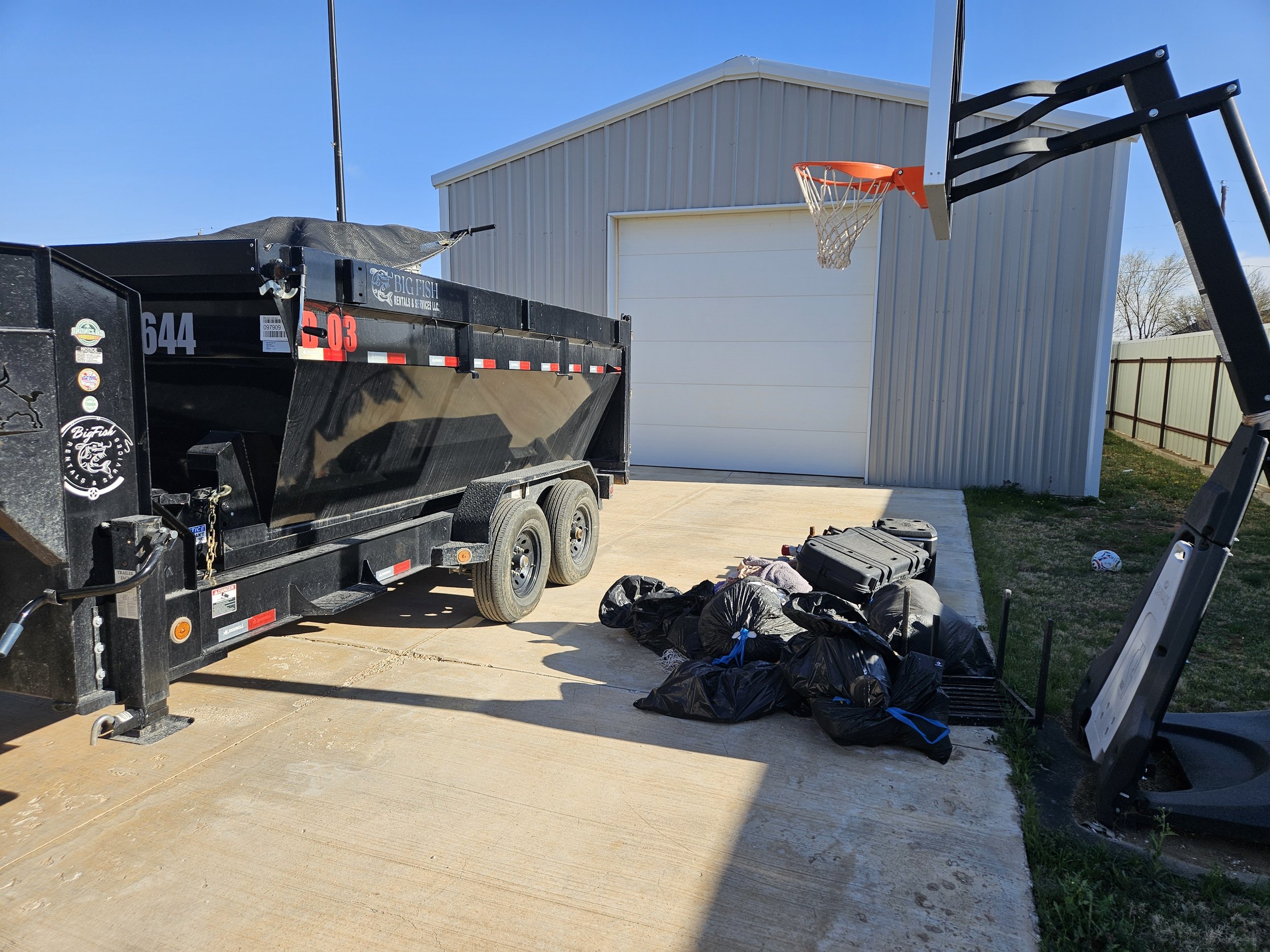 A trailer parked on a concrete driveway, with a basketball hoop nearby. There are black garbage bags and some tools on the ground in front of the trailer, and a building with a white garage door and a metal fence in the background.