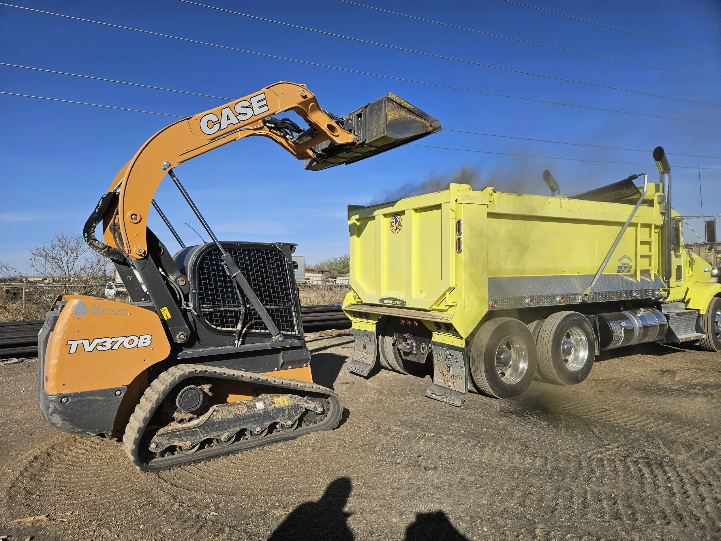Construction equipment on a dirt lot, including a small excavator labeled 'CASE' and a large yellow dump truck with a black container and mud on its tires, under a clear blue sky with power lines overhead.