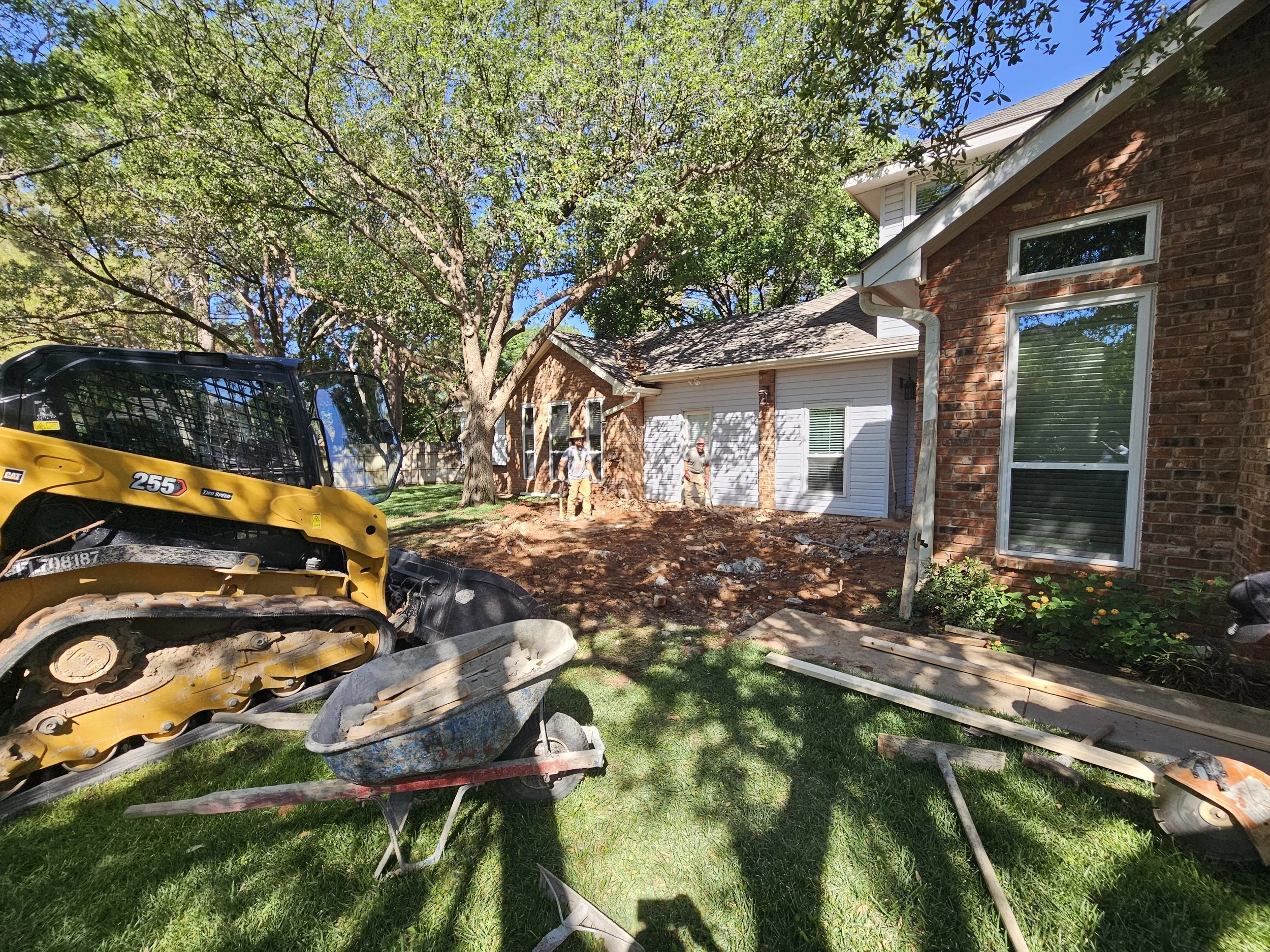 Construction workers working on the exterior of a brick house with trees, construction equipment, and tools in the yard.