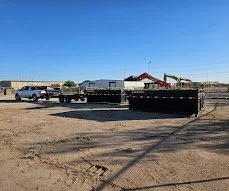 Construction site with a flatbed trailer on dirt ground and several vehicles parked in the background under a clear blue sky.