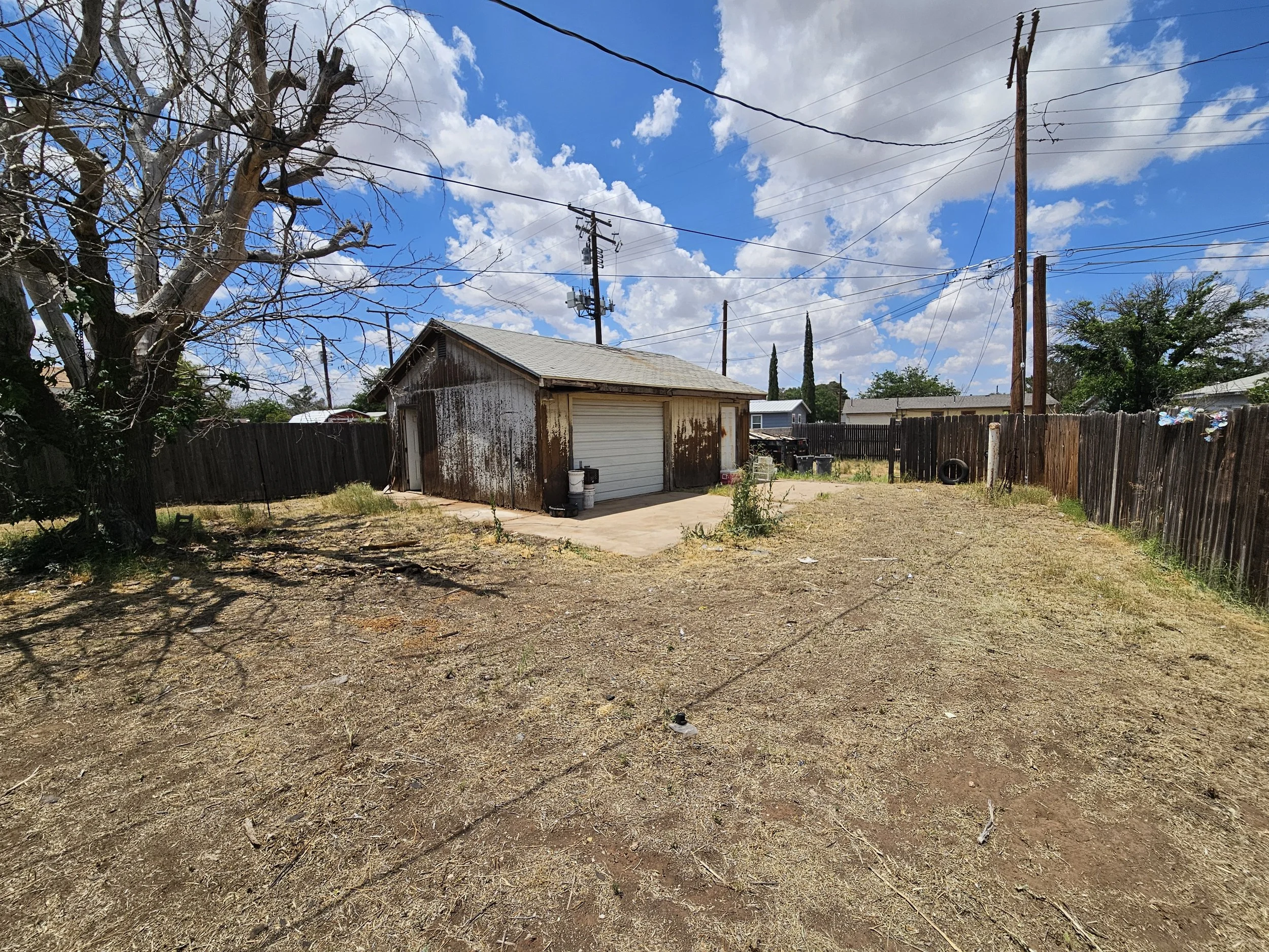 A backyard scene with a large, leafless tree on the left, a small, weathered shed with a garage door in the center, and a wooden fence on the right. Overhead, power lines cross a partly cloudy blue sky. The yard has patches of grass and dirt, with so