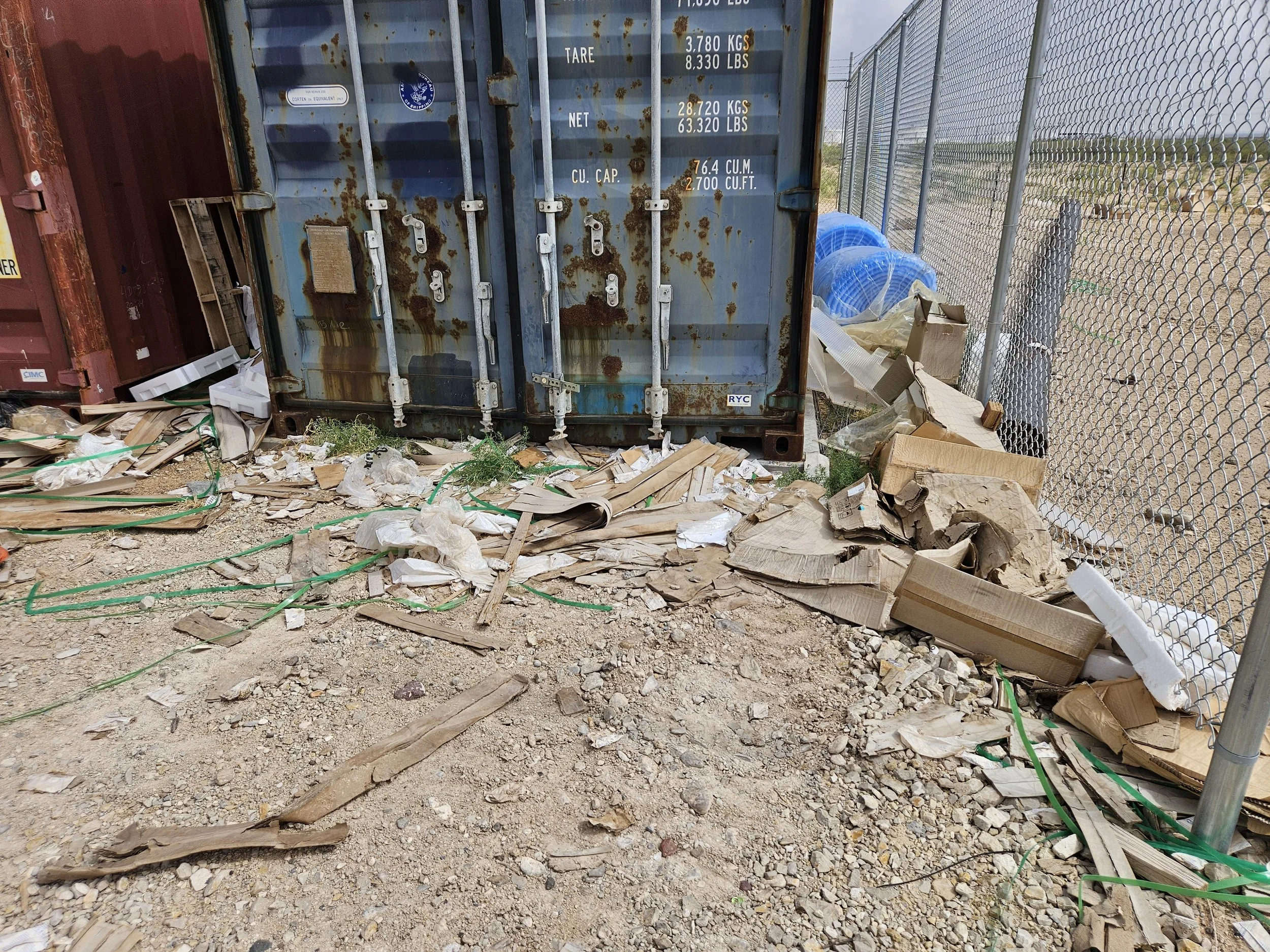 Rusty blue shipping container behind a chain-link fence, surrounded by scattered cardboard boxes, wooden planks, and debris on sandy ground.