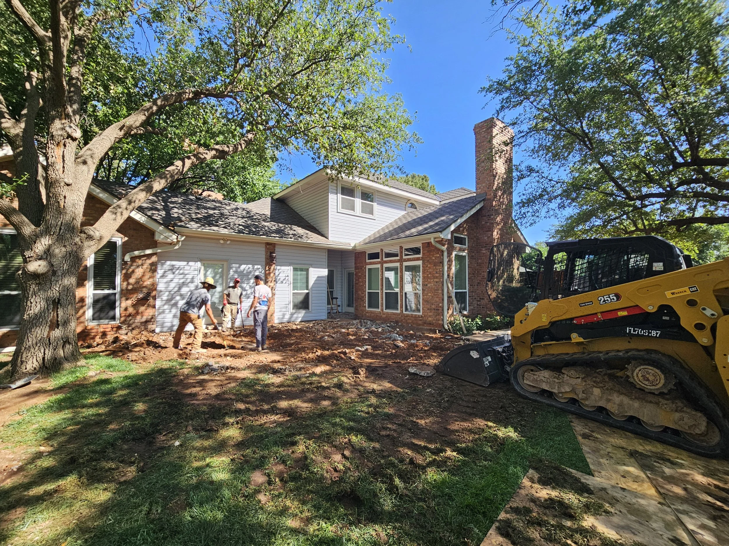 Workers are landscaping and working on soil in the backyard of a house with trees and a chimney, operating a small tracked construction vehicle.