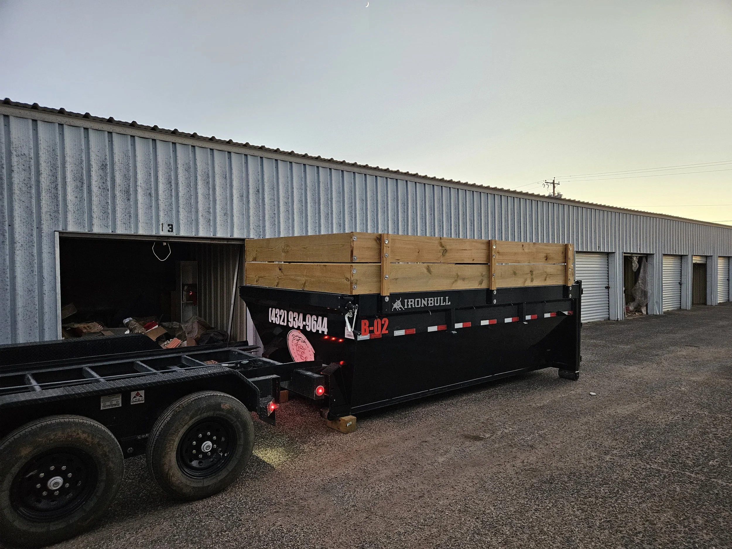 An industrial building with a corrugated metal facade and a partially open door. In front of the building, there's a black trailer with a wooden railing on top and visible tires, parked on gravel ground during dusk or dawn.