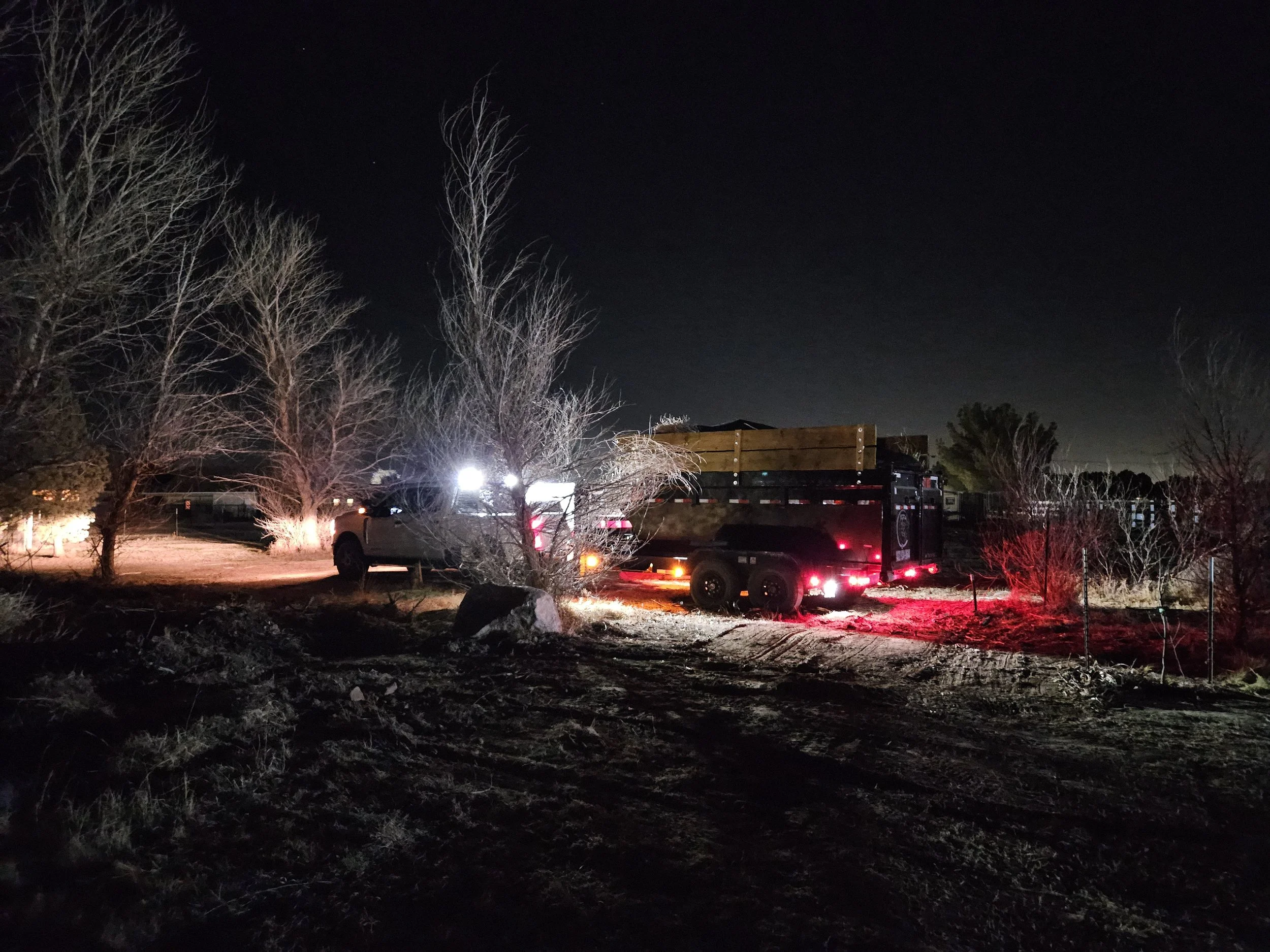 Nighttime scene with leafless trees, a pickup truck with a trailer and bright headlights, and a dark sky.