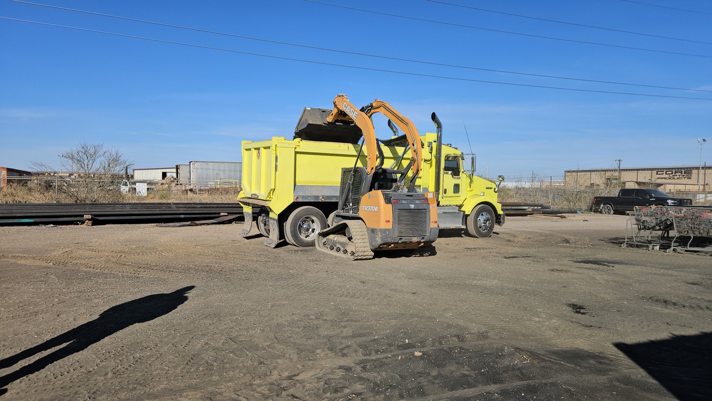 A construction lot with a yellow dump truck and excavator, a black pickup truck, shopping carts, and industrial buildings in the background under a clear blue sky.