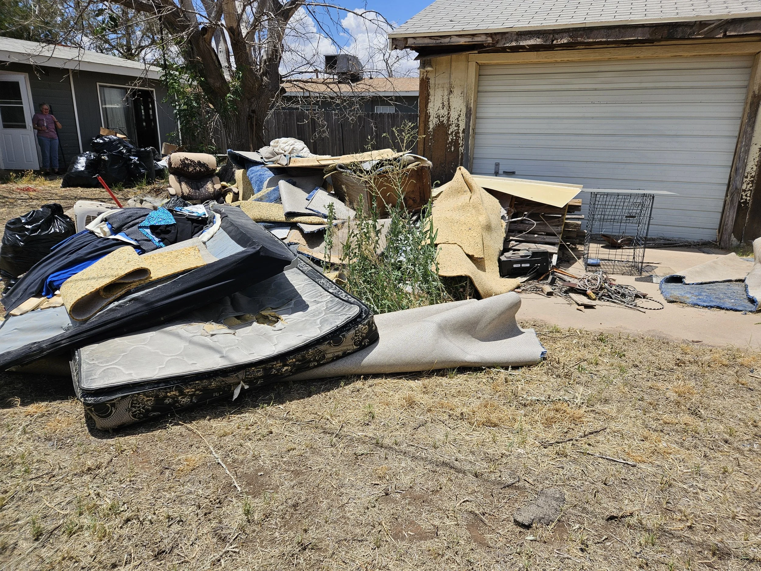 Backyard with pile of discarded furniture, mattresses, and trash next to a house with gray siding. A person is standing near a door, and a tree with bare branches is in the background.