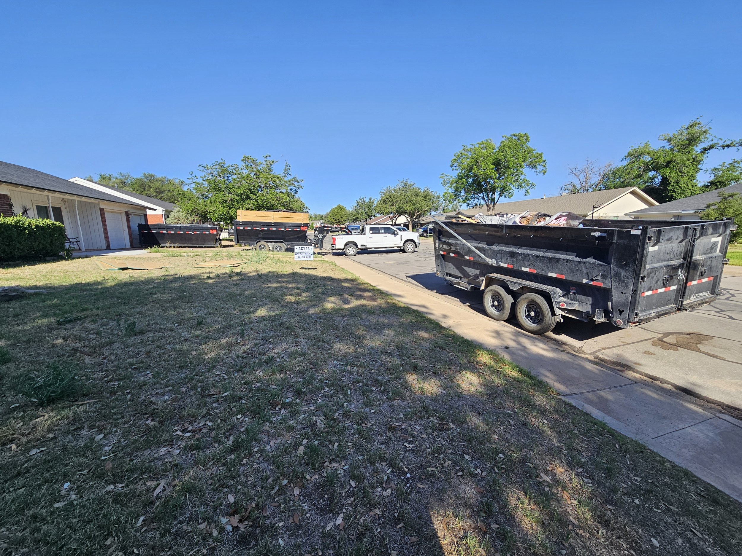 A residential street with multiple trash dumpsters filled with debris. There are several houses with trees in the background under a clear blue sky.