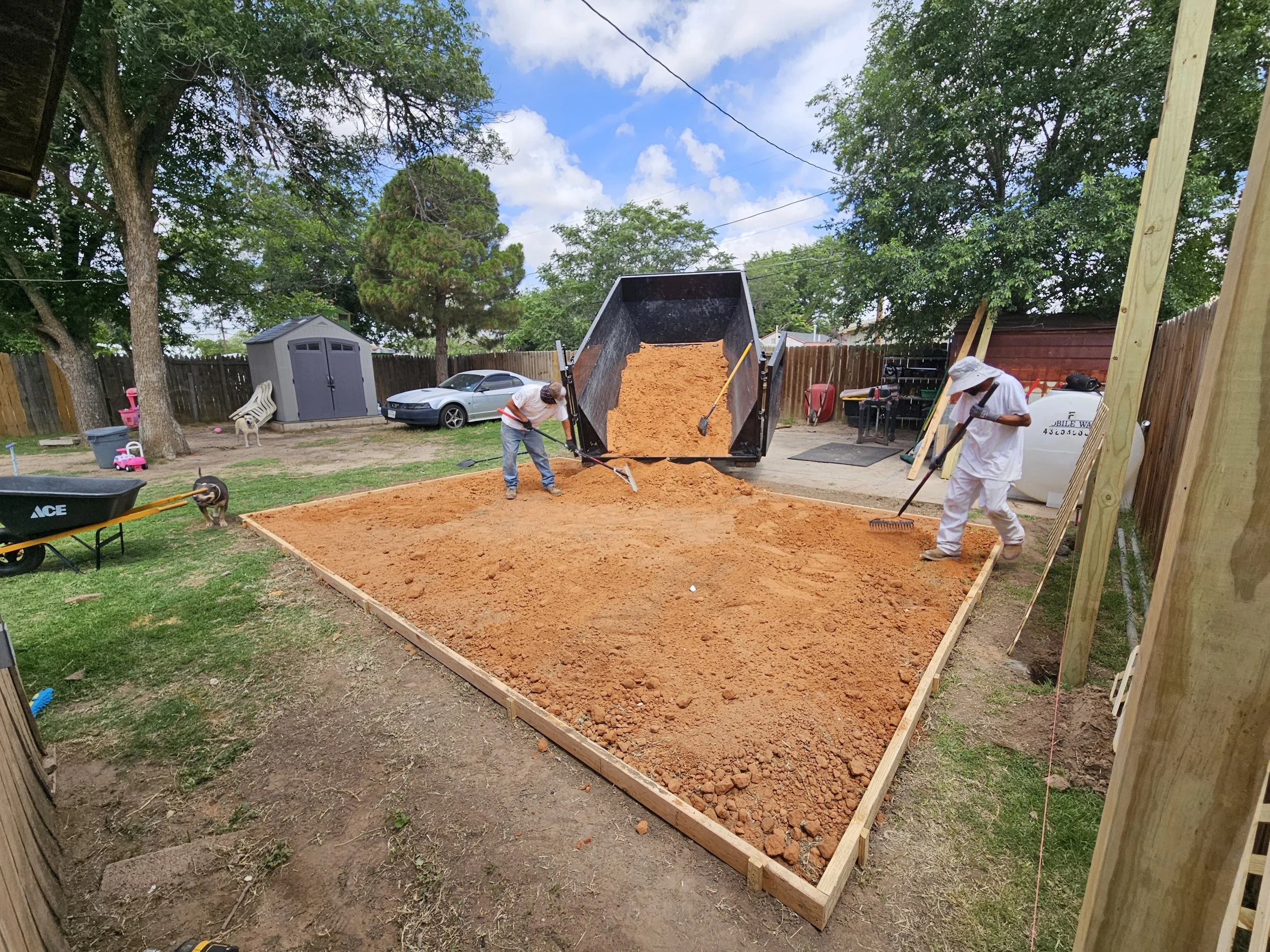 Two workers are leveling dirt in a backyard frame for a construction project, with a dump truck emptying dirt nearby. There are trees, a shed, a car, and various tools and supplies around.