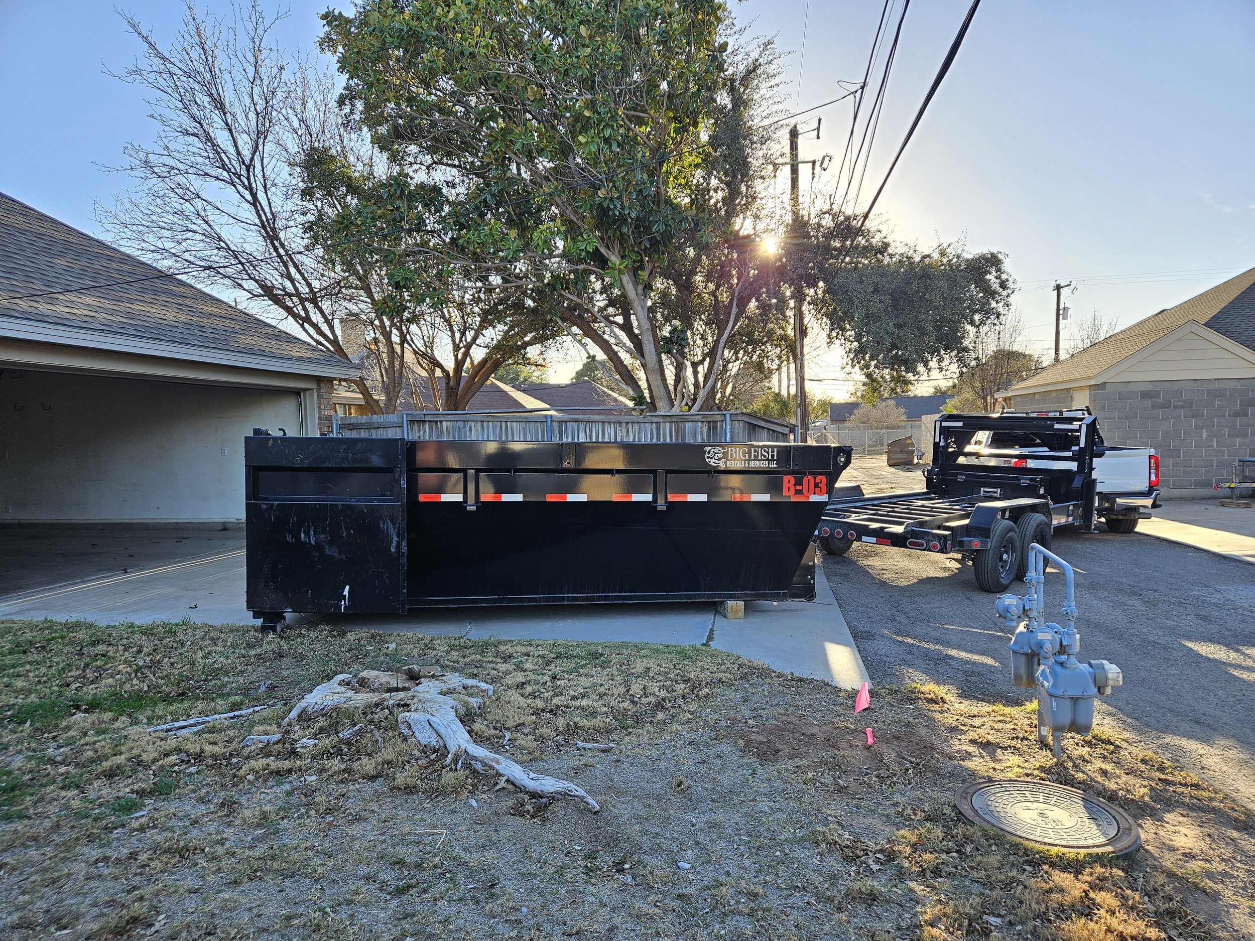 A backyard scene at sunset with two large trees, a chain-link fence, a building with a shingled roof, a utility truck and trailer, a fire hydrant, and some patches of grass and dirt.