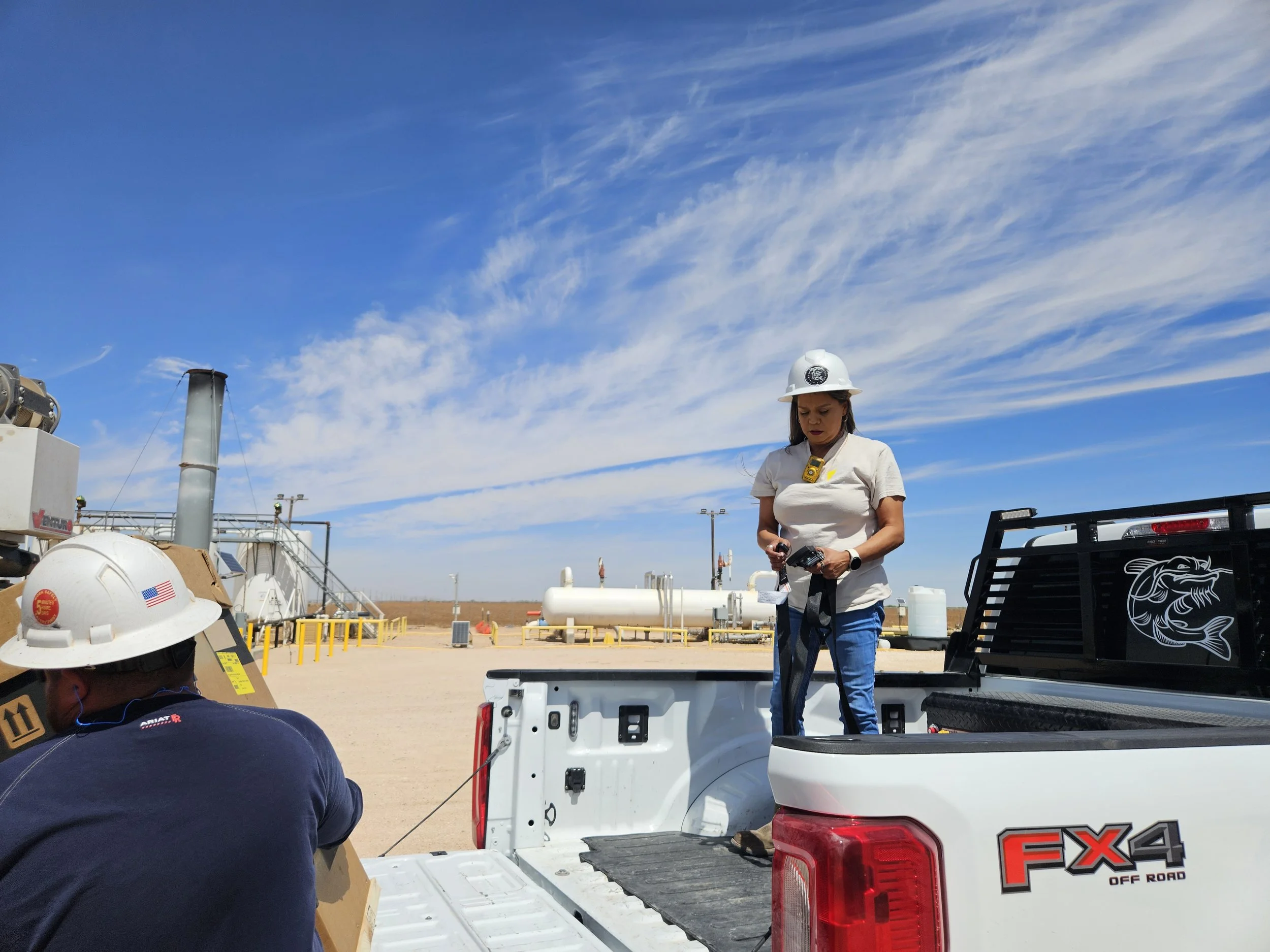 A woman wearing a white hat and beige shirt is standing at the back of a white pickup truck with an open tailgate, working with tools. Other workers are visible in the foreground and background, with clear skies and industrial equipment around.
